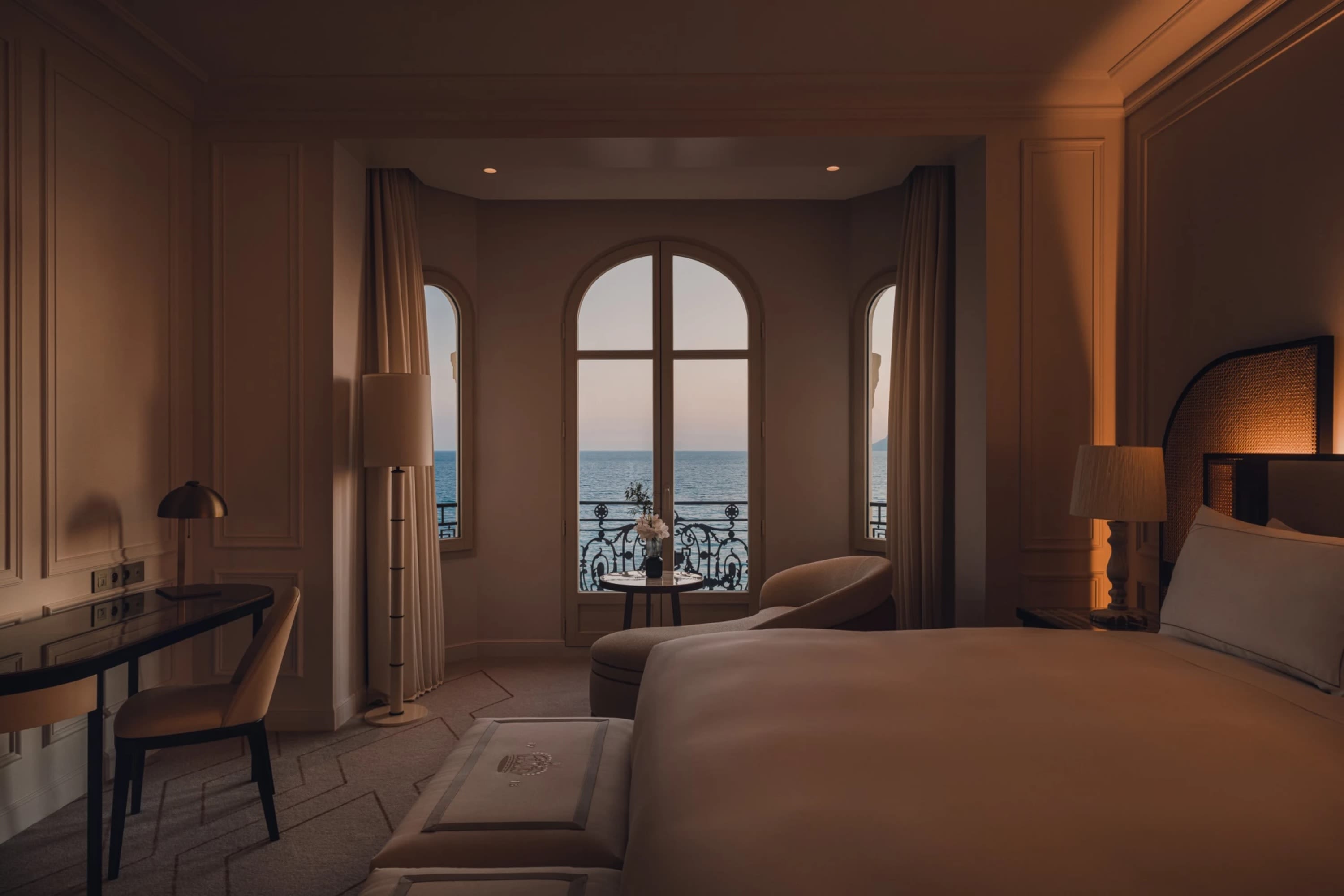 Moody-lit room with high-end, classic furnishings and ornate windows overlooking calm ocean waters
