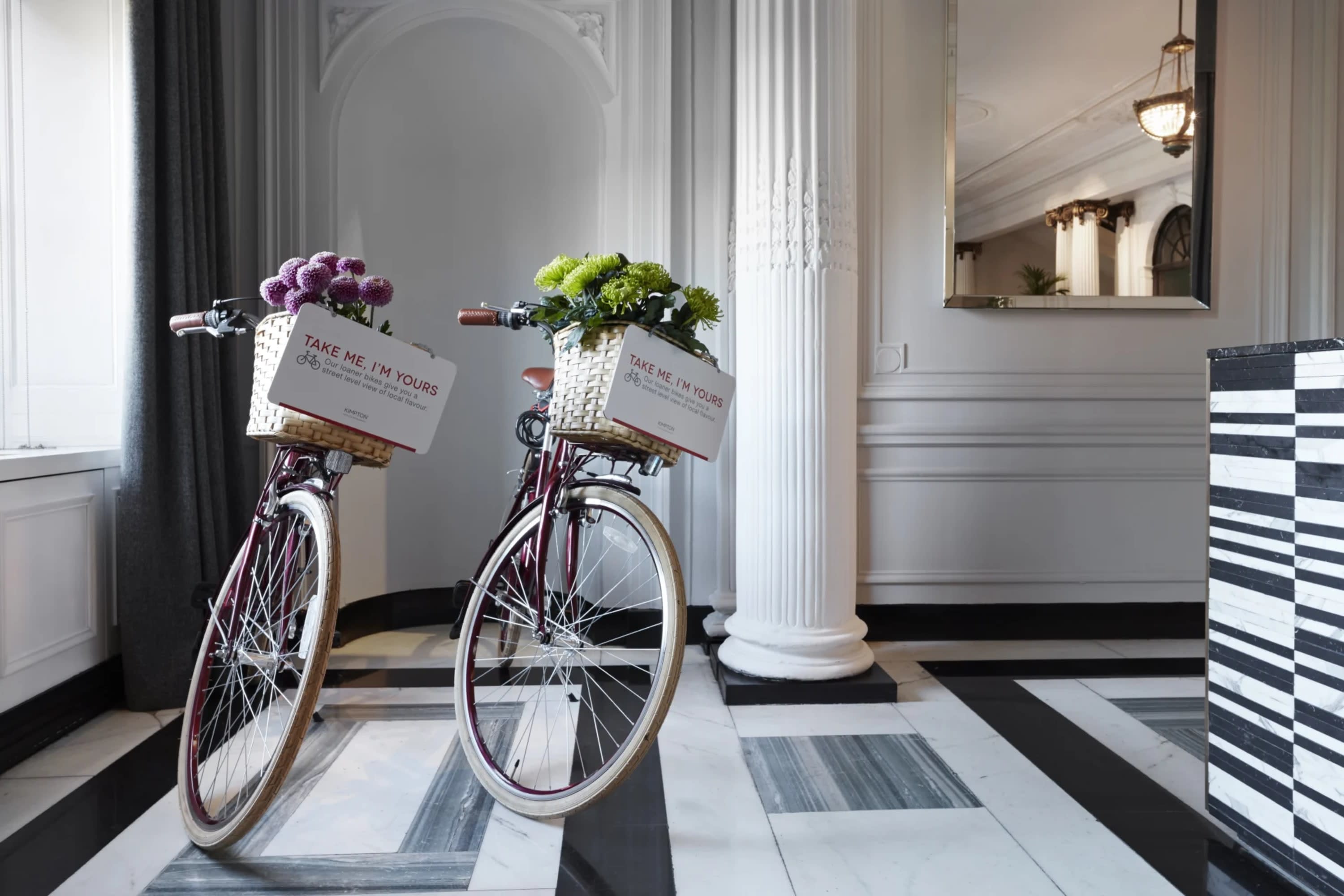 Two beachcruiser bikes stand next to each other, floral arrangements in their baskets, on marble floors next to a whitewashed classical column