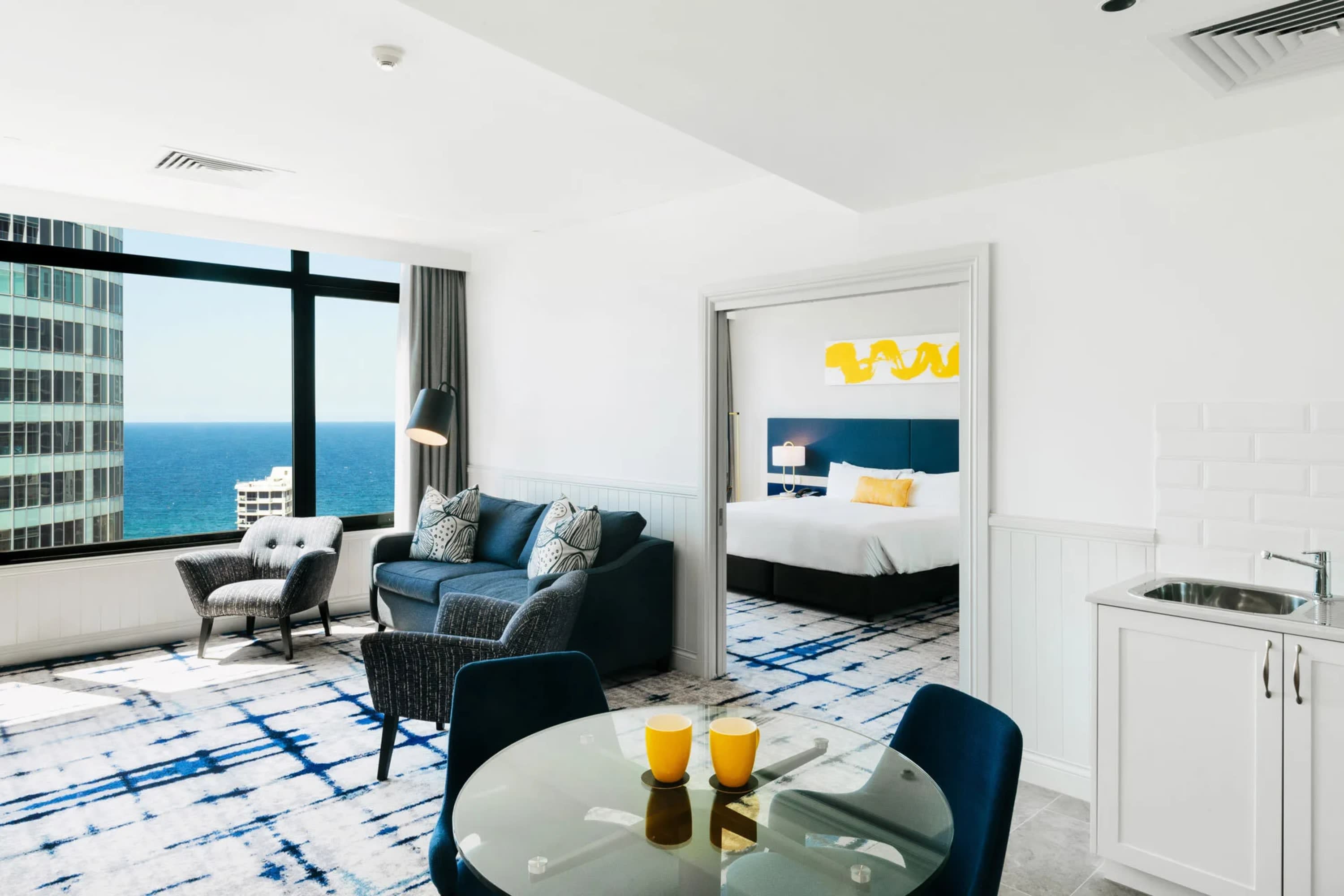 Trendy decor with a blue and gold color palette in a spacious, whitewashed living room. A large primary bedroom is visible through an open doorway and the ocean is visible through large windows