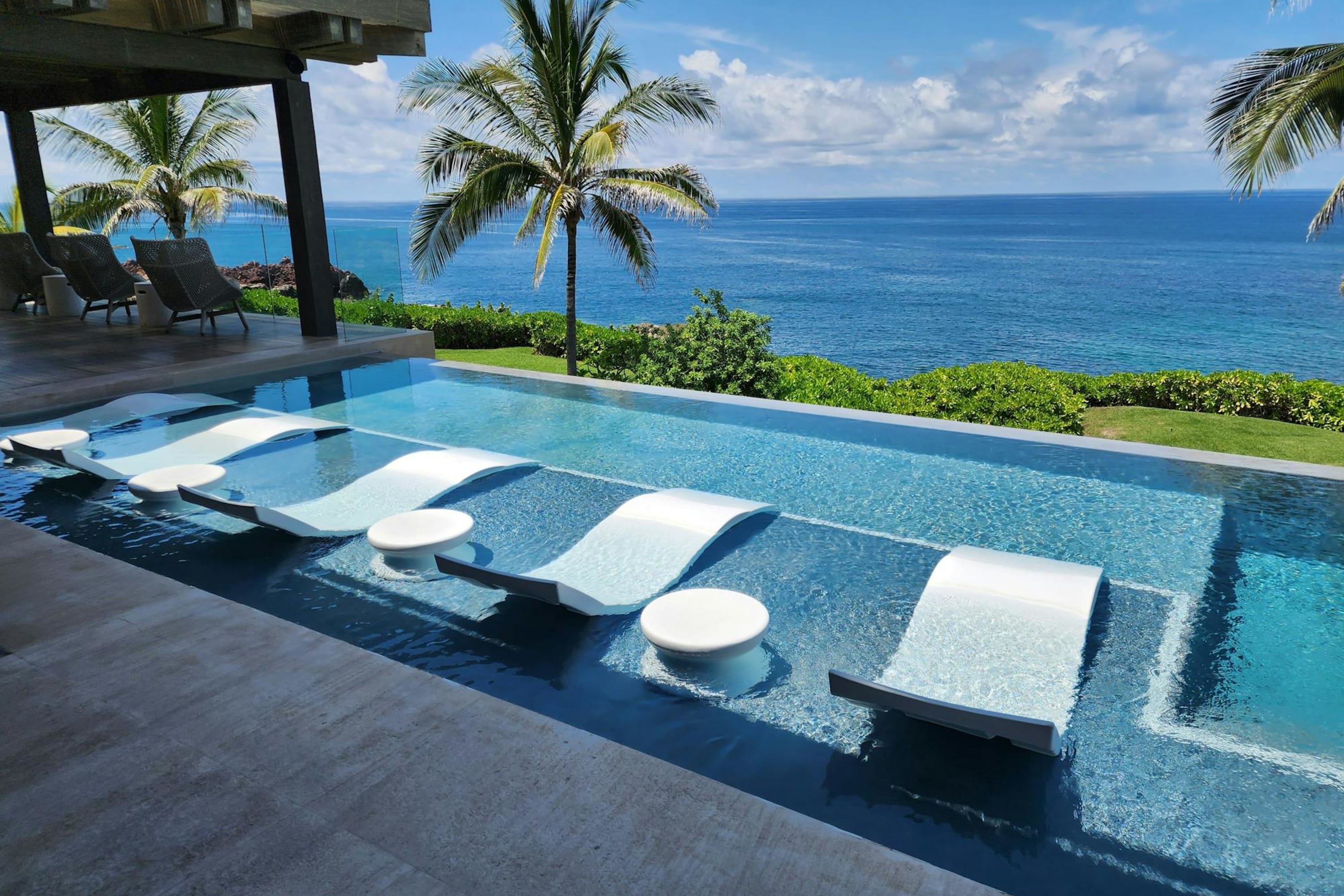 Chic white loungers and small tables sit directly in the water of a pool with an ocean view and palms