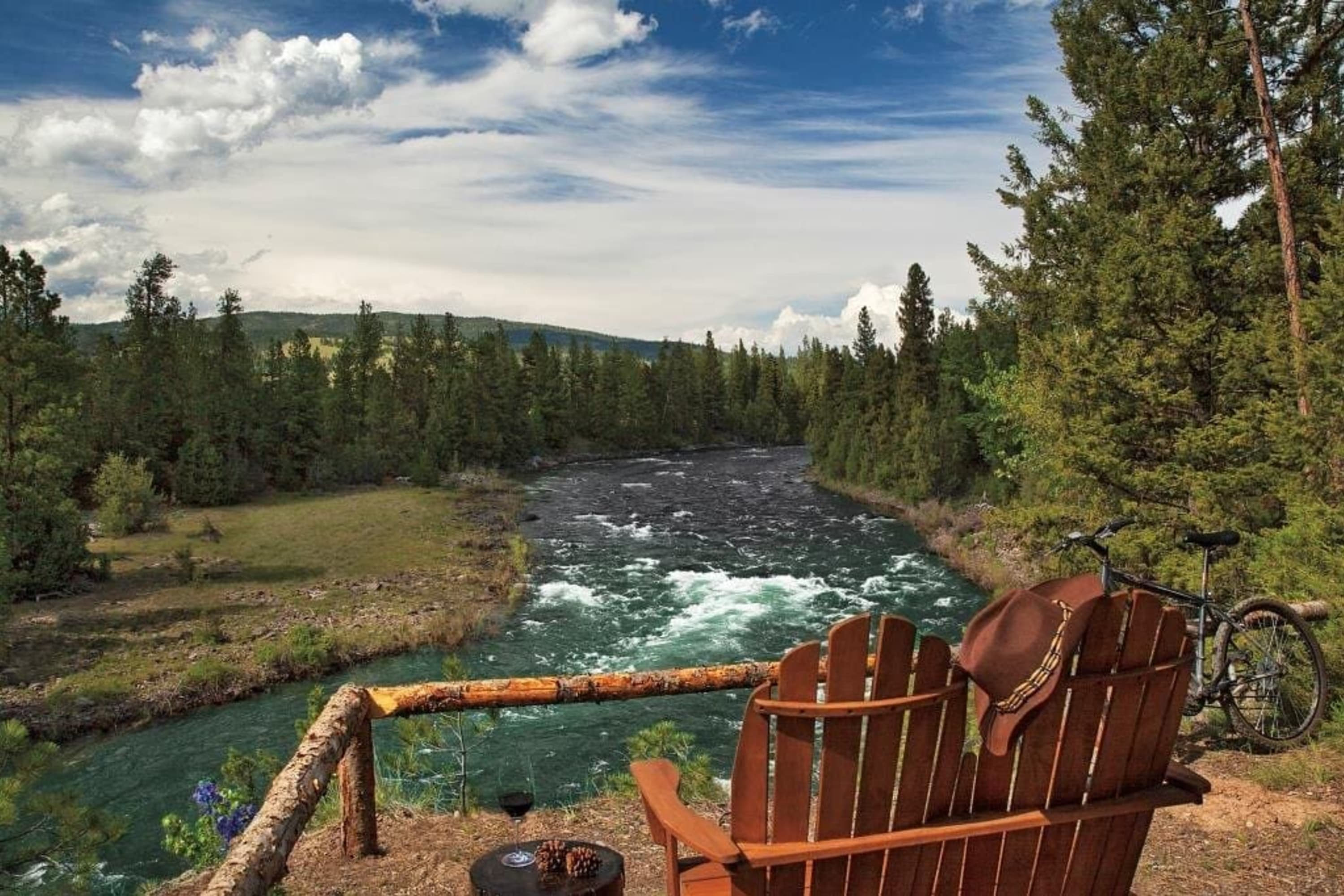 Rustic fencing and a pair of old wooden chairs are all the separates the camera from stunning blue river rapids and verdant pines