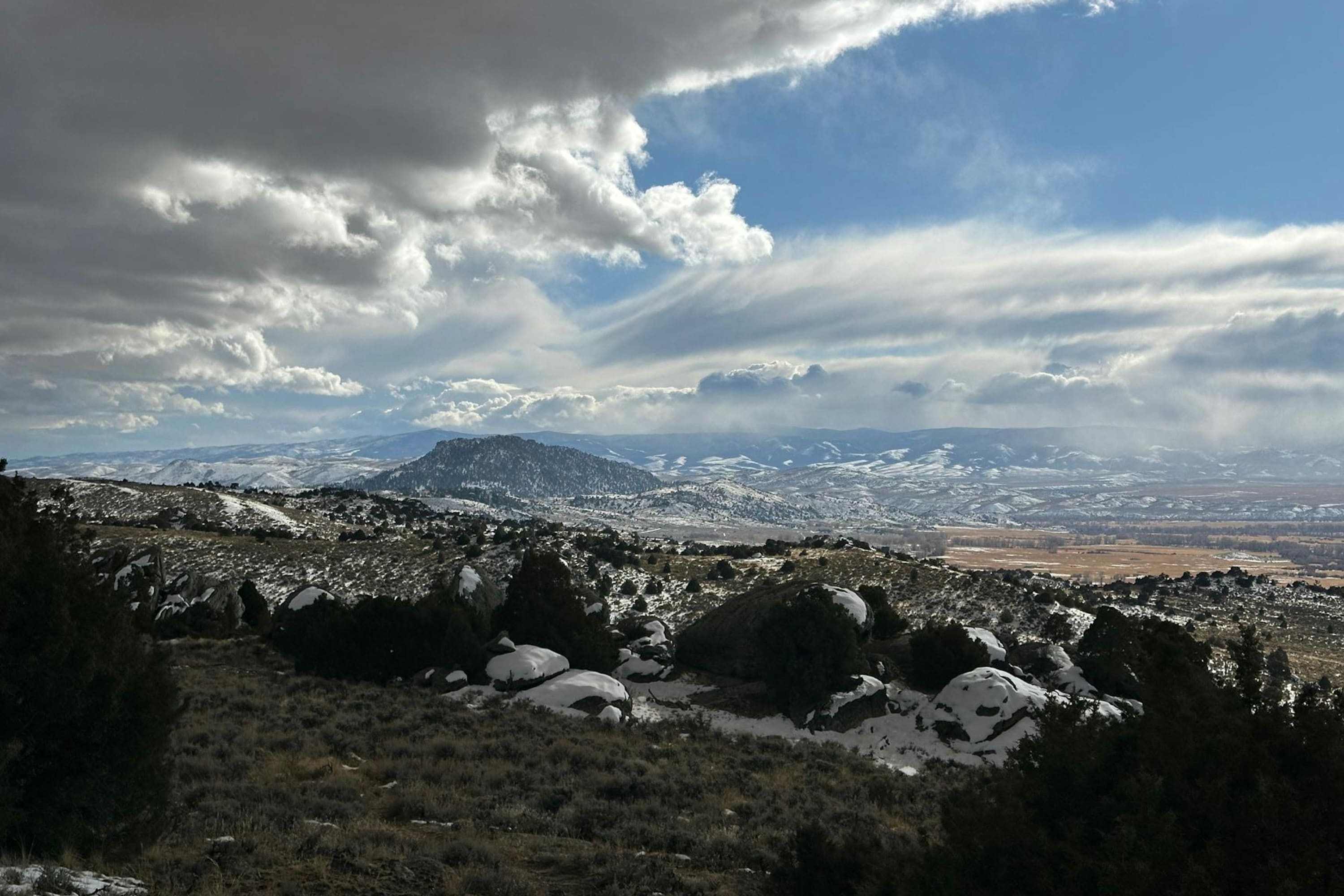 Snow sparsely blankets an untamed landscape interrupted by sudden mountain ranges and pine forests under massive clouds