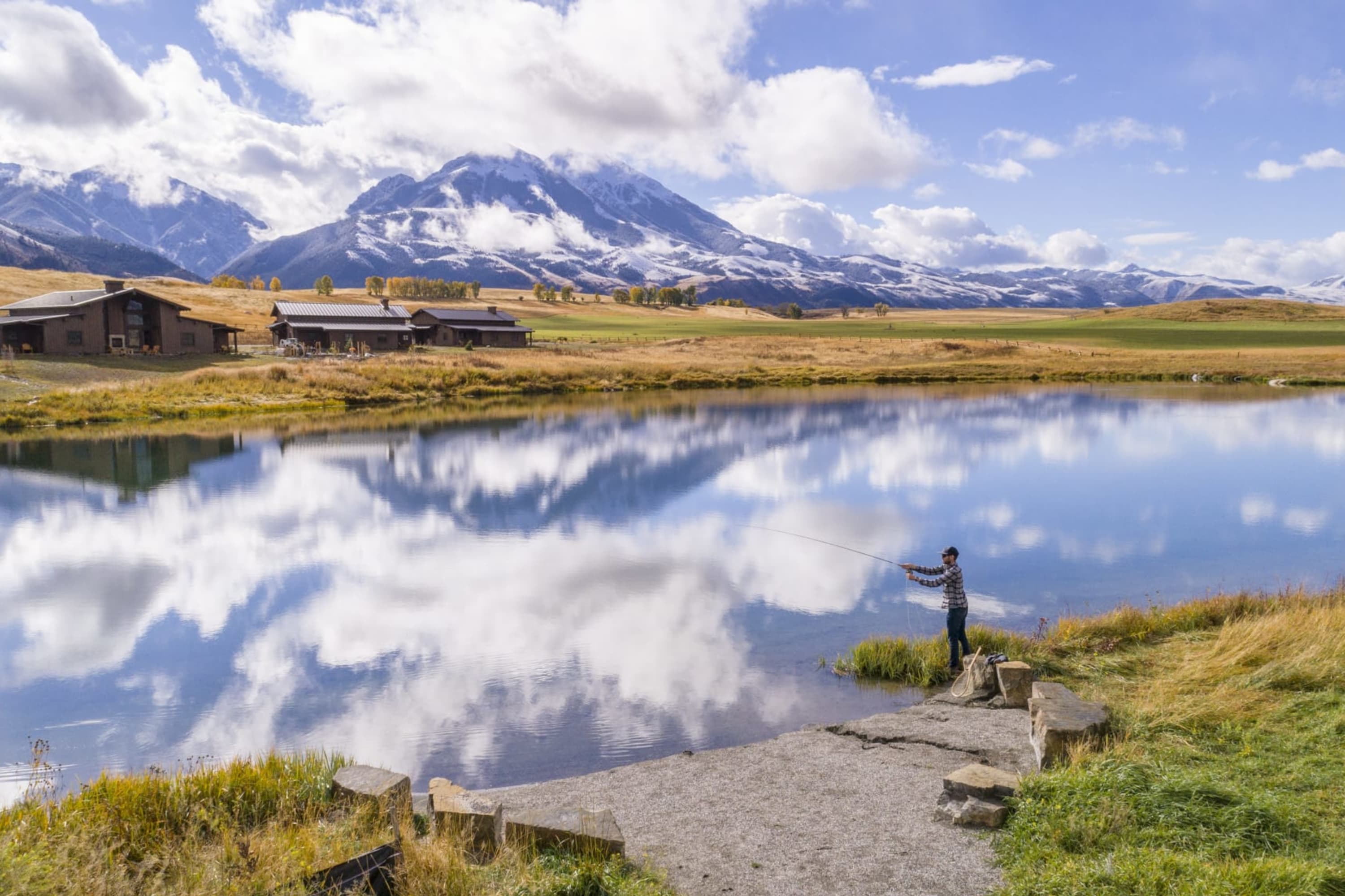 A lone man casts a line into a large body of water that's reflecting a mirror image of a wide open, somewhat cloudy sky. In the background, prairies extend for miles until a snowy mountain