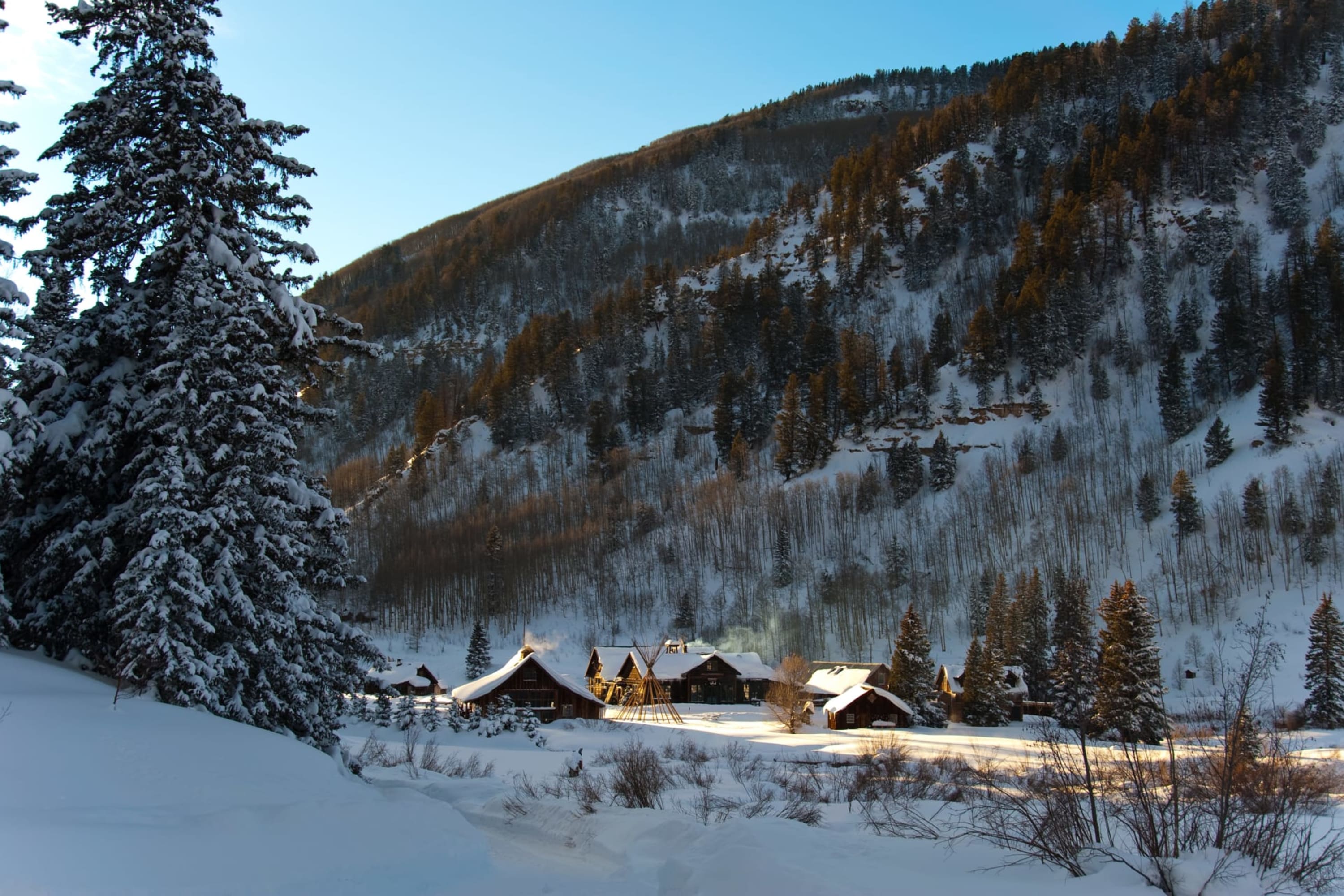 What appears to be a small town of old log cabins sits at the bottom of a valley. A snowy tree frames the photo and a snowy mountain stands in the distance