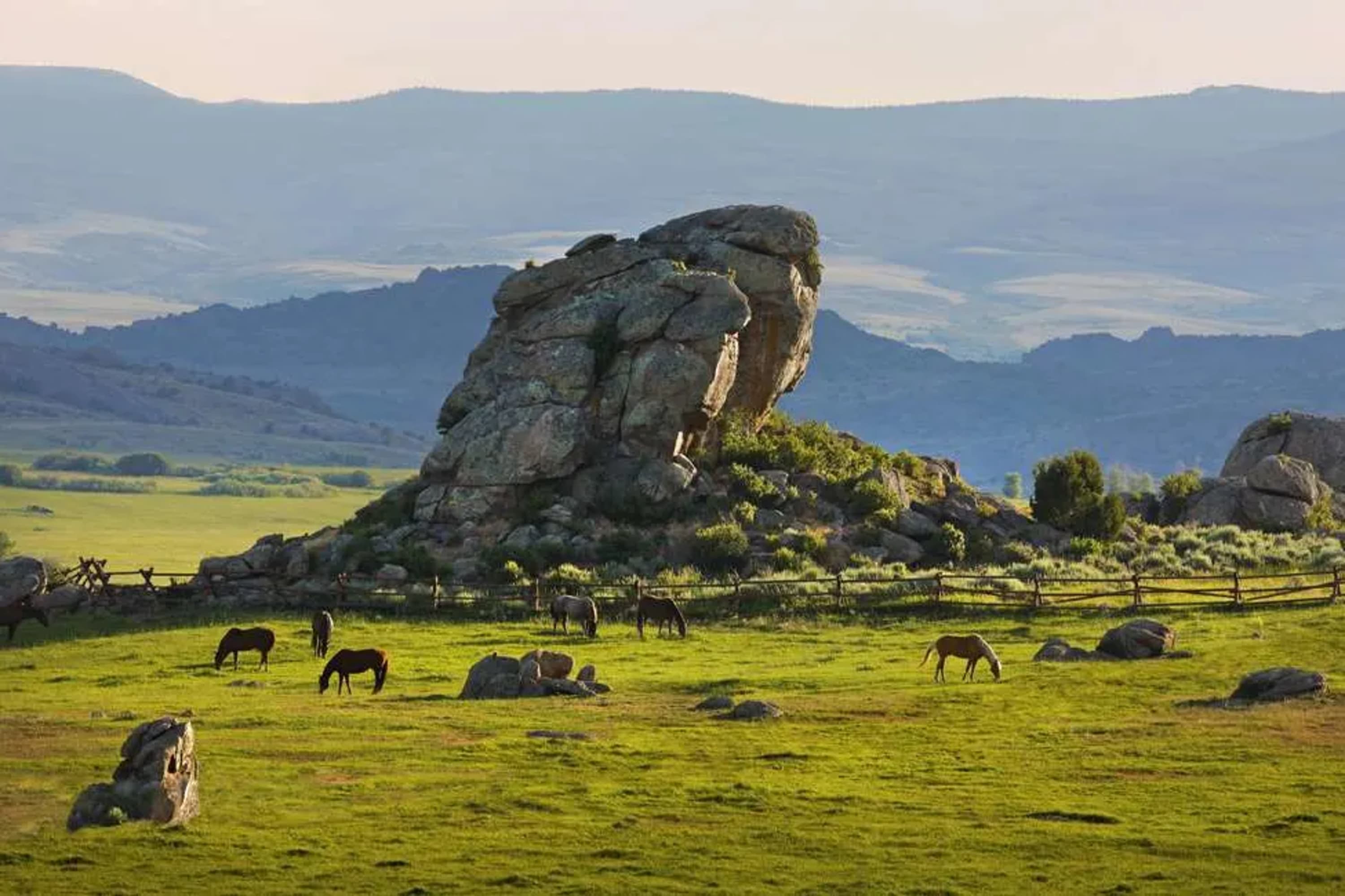 A few horses graze on green grass amid rocky outcroppings. Hills and mountains rise and fall in the distance
