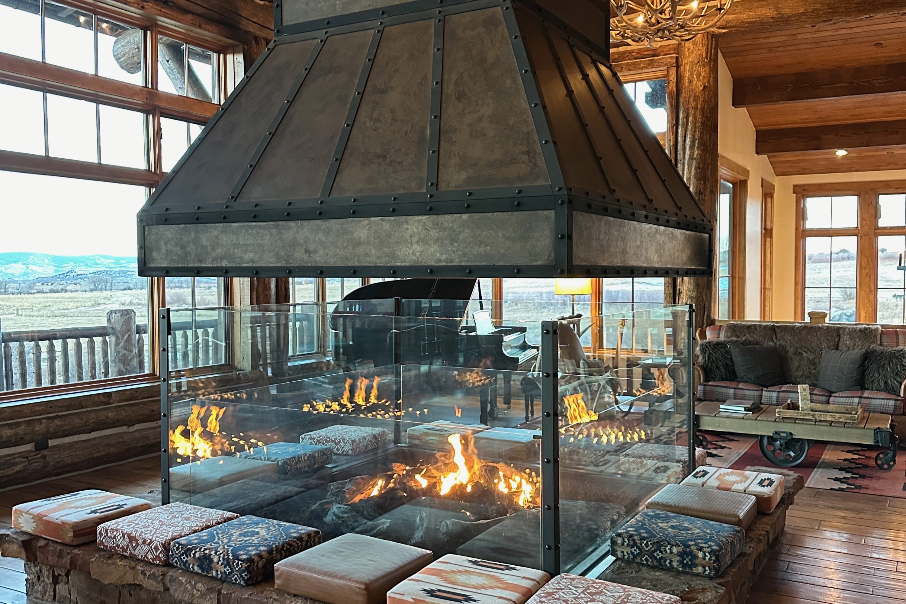 A massive gas fireplace lit up within a lodge-like building. A grand piano, elegant furniture and stellar views are visible behind the fireplace's glass walls