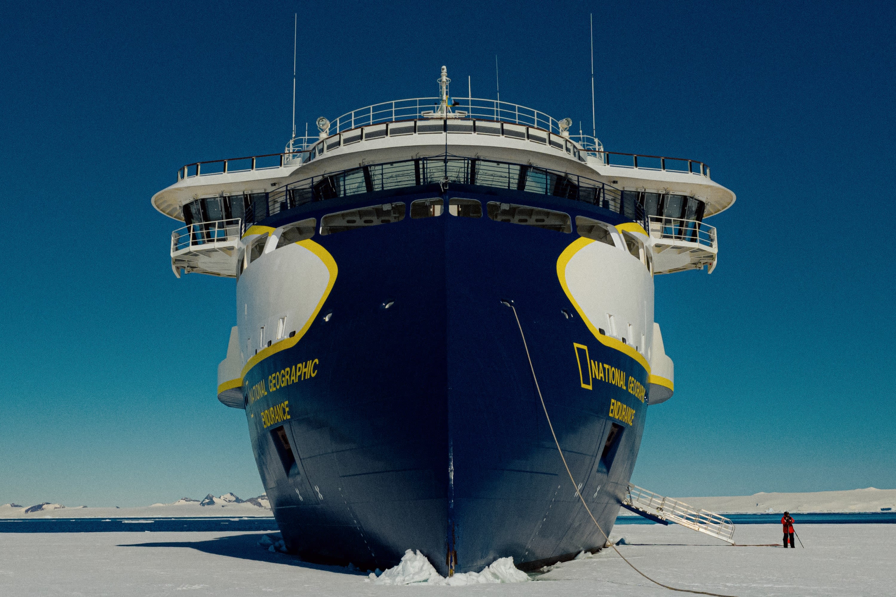 A massive icebreaker cruise ship 'beached' on the ice in Antarctica. A person is standing next to the ship to provide scale