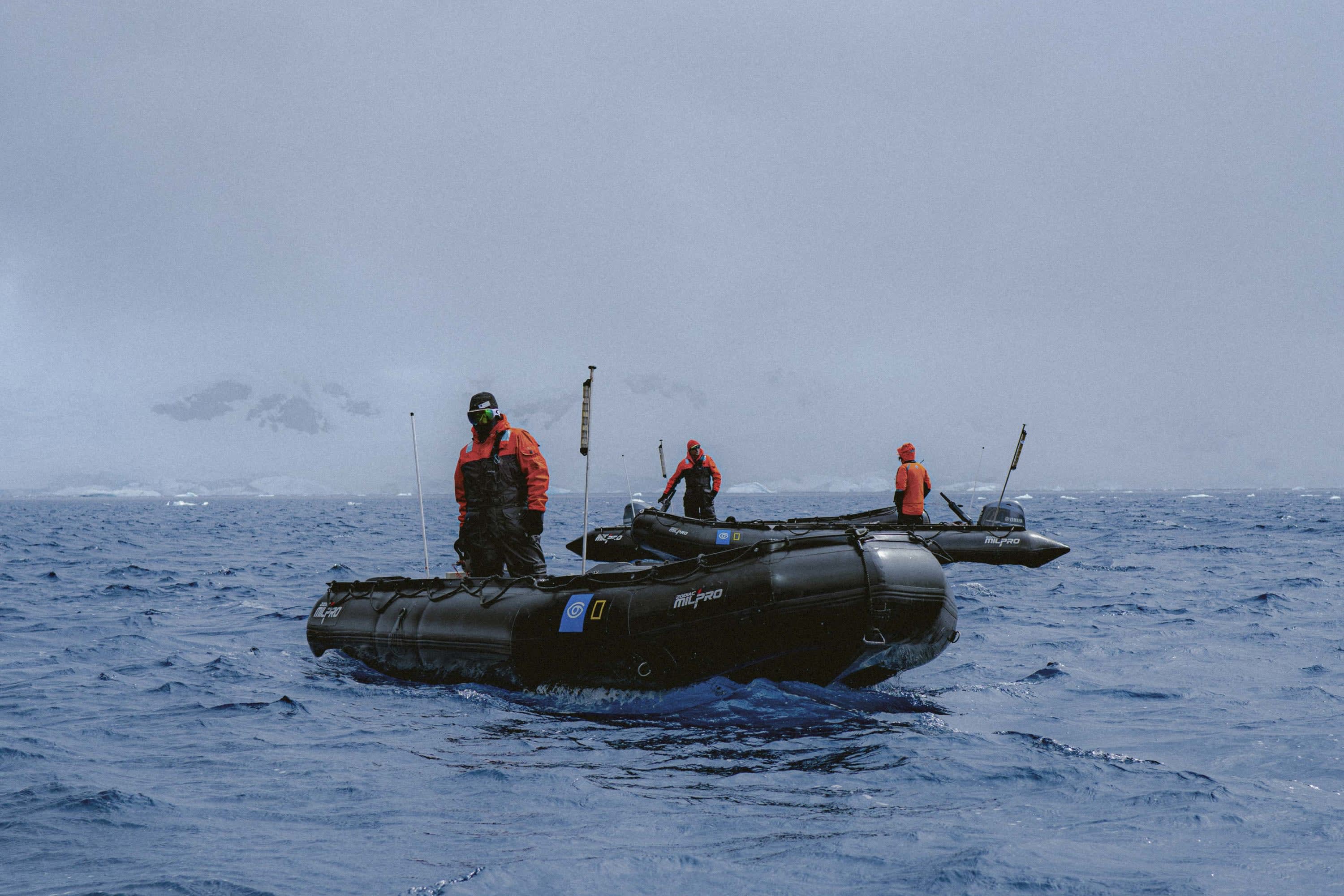 A few crewmembers in orange and black winter clothing on two zodiac craft in calm, cold waters