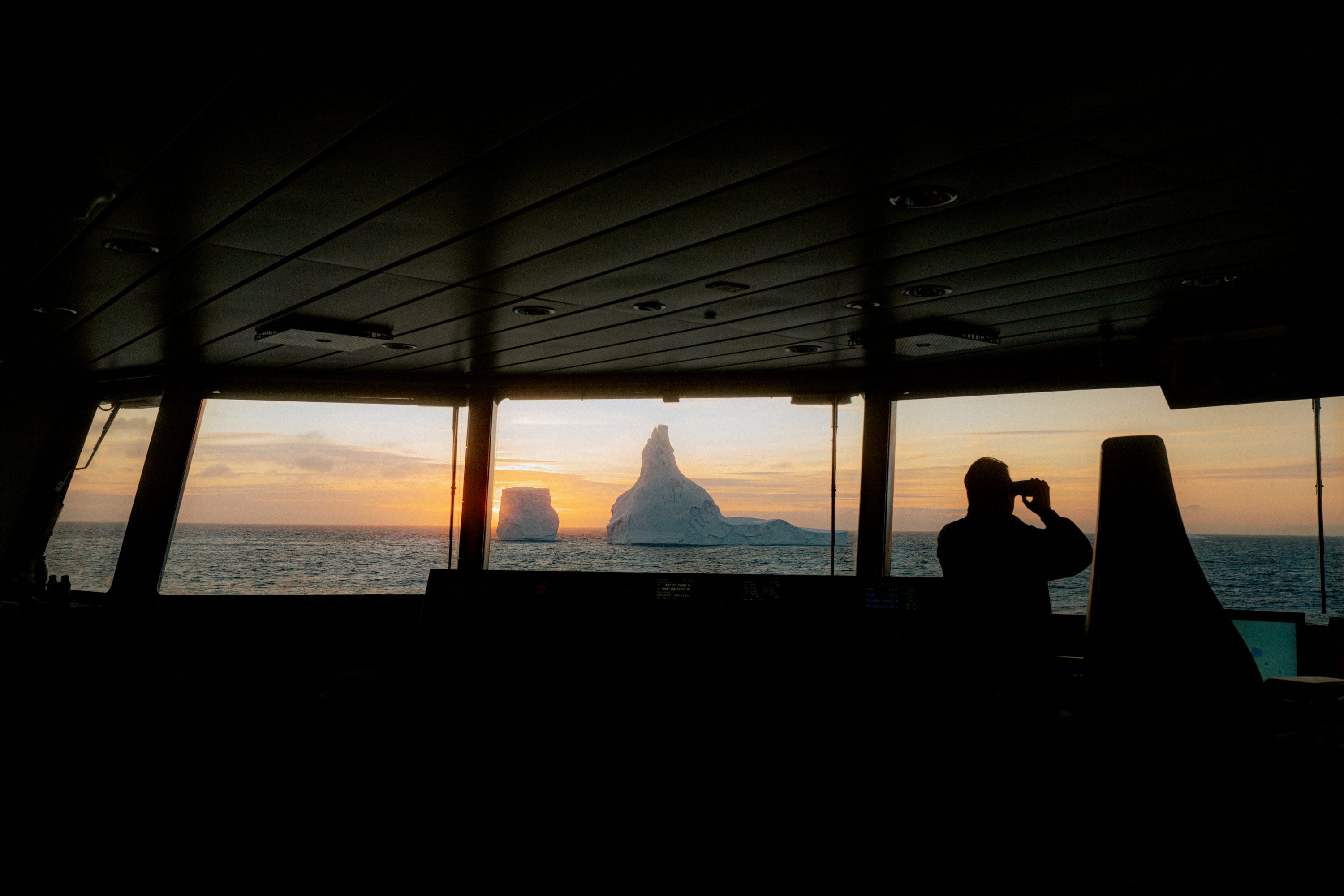 A crewmember looks out of a viewport with binoculars with icebergs in the distance