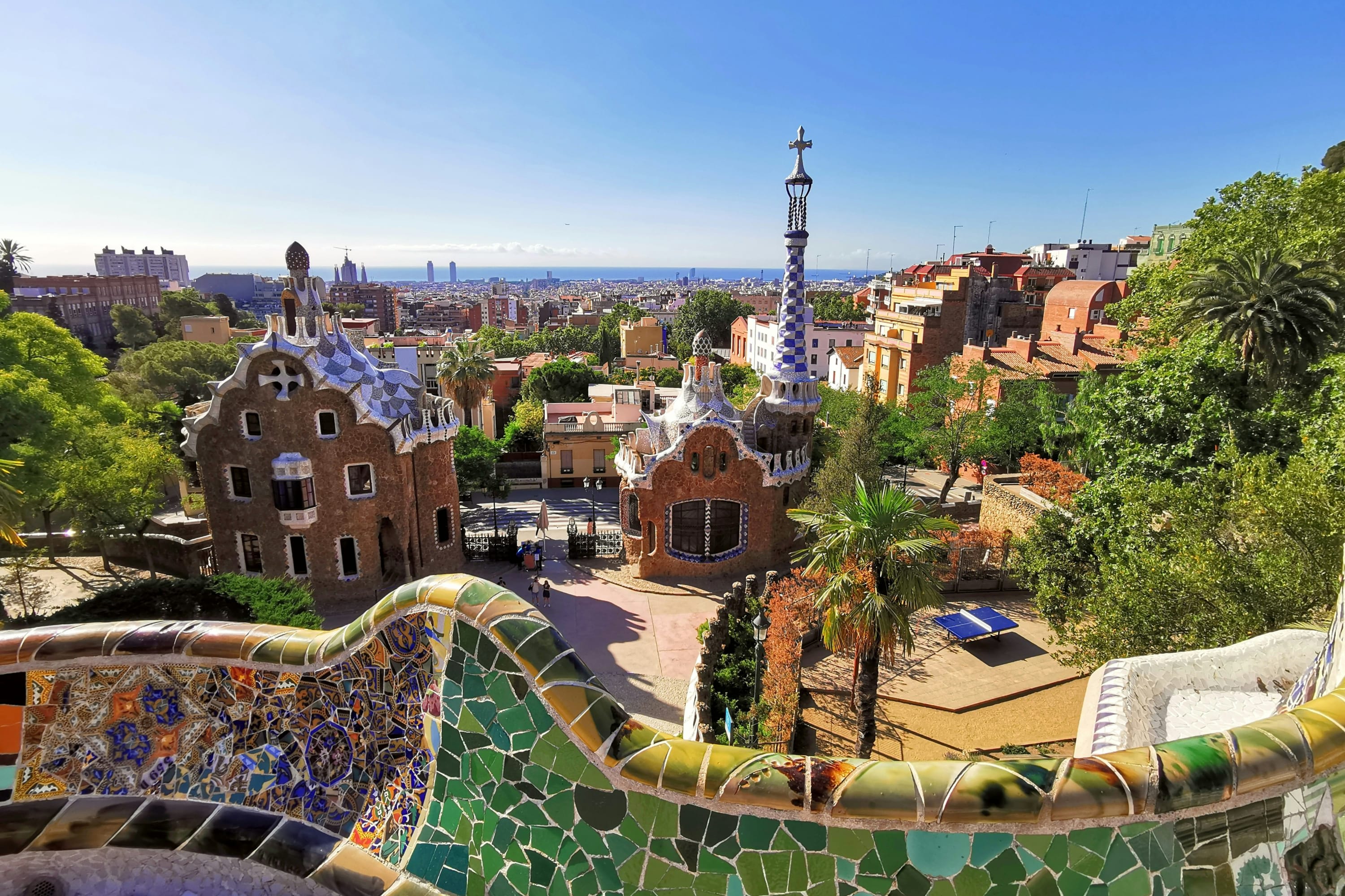 A stone fence with the tilework of Antoni Gaudi partially obscures a skyline view of Barcelona, with the Mediterranean faintly visible in the distance