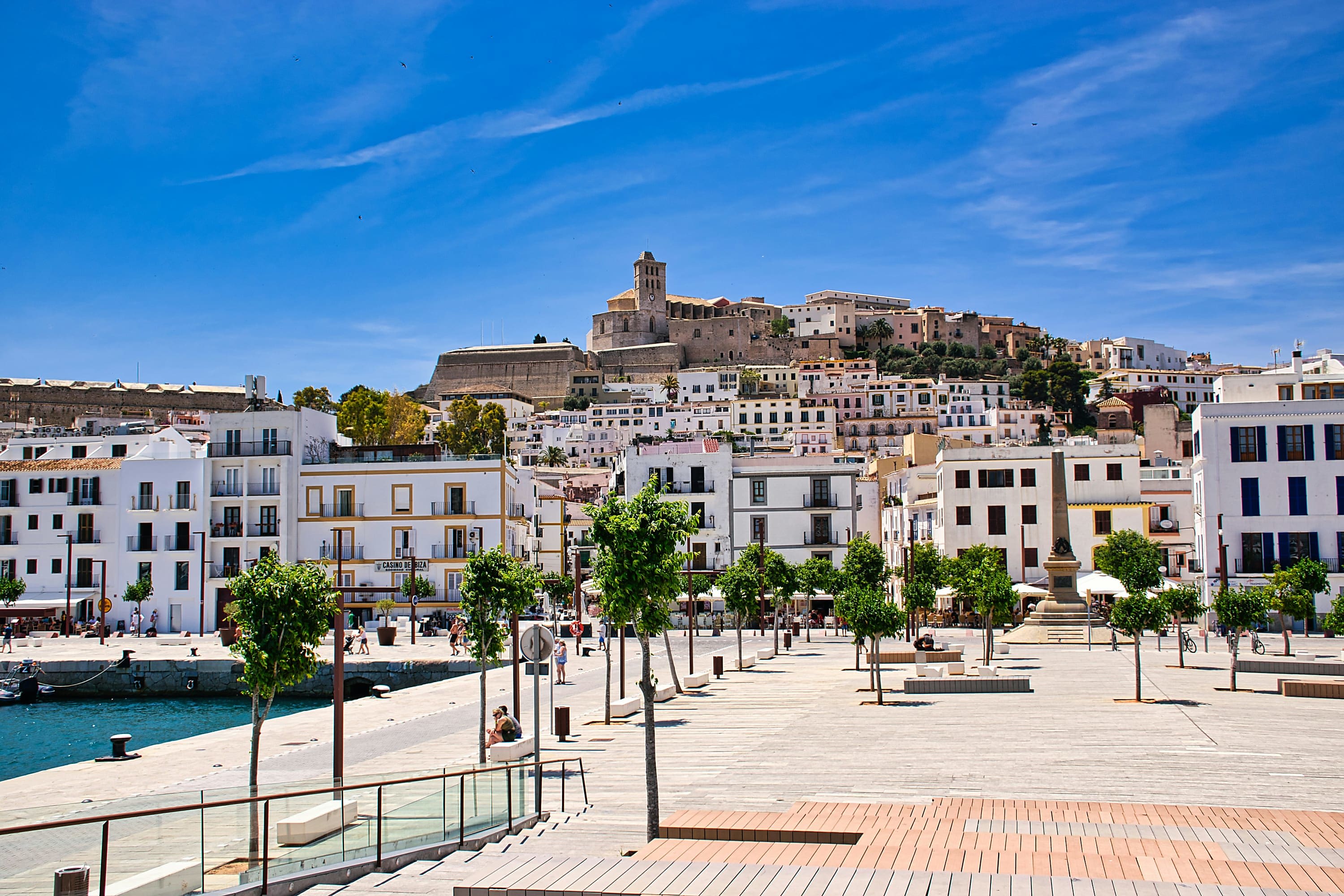 Whitewashed historical buildings with large dark windows at the end of concrete pier. In the distance, an imposing old fortress stands atop a hill