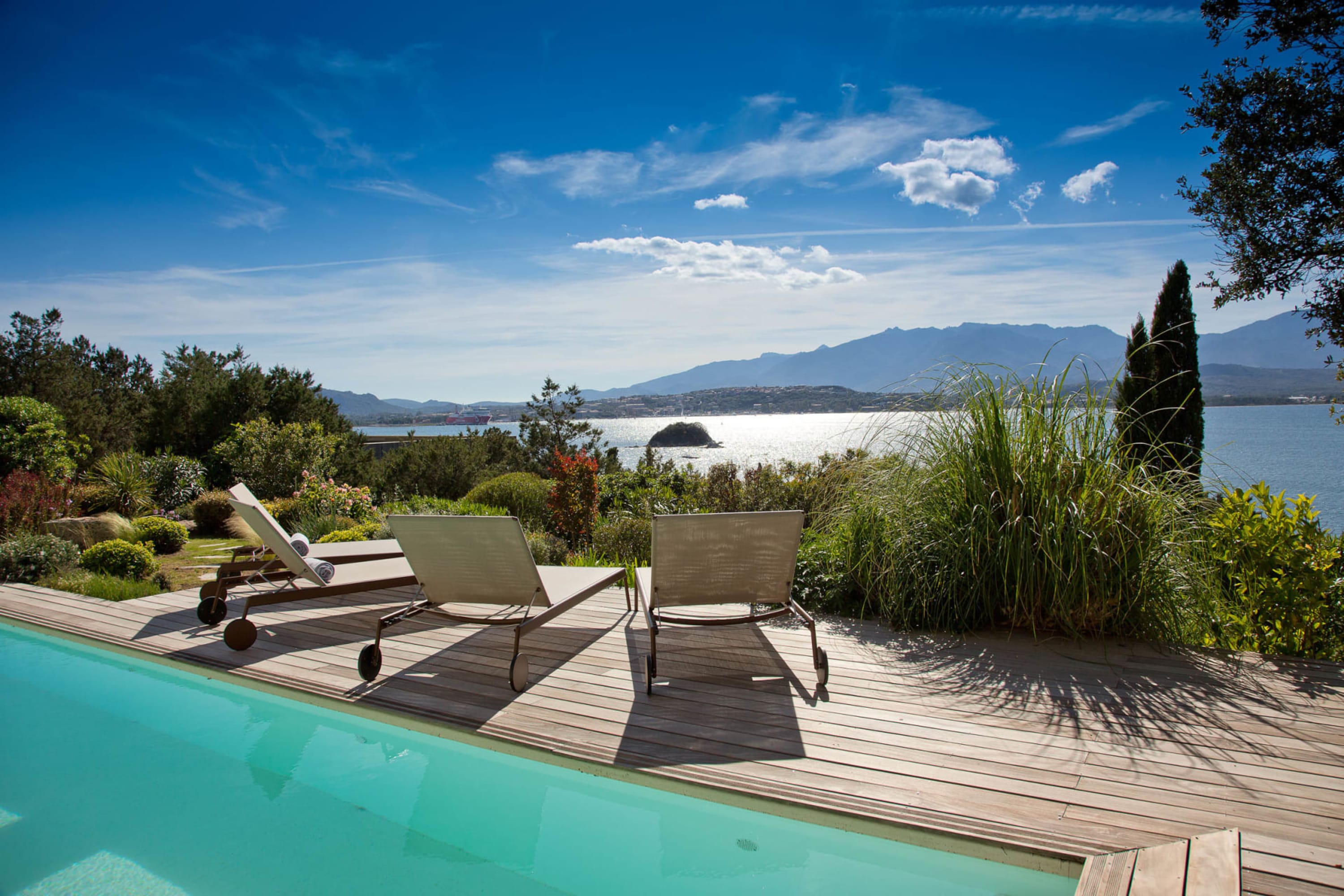 A pool is separated by a wooden patio with loungers and a small garden space from the Medeterranean