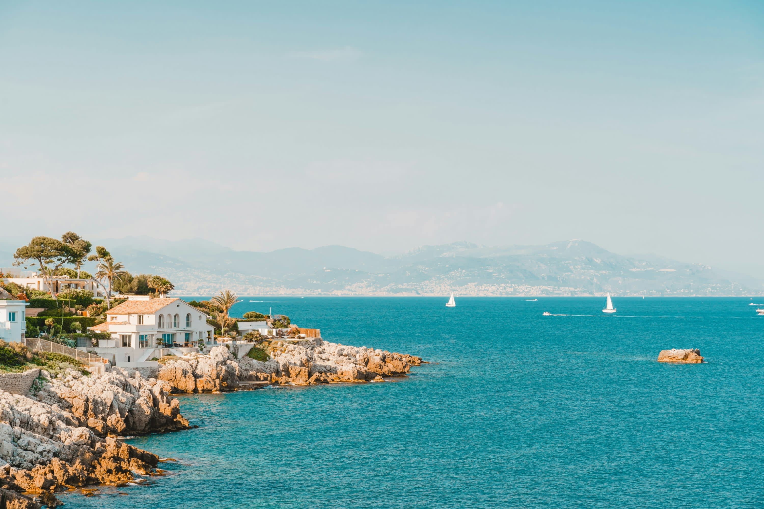 A rocky coastline with a Mediterranean-style house and turquoise-blue water with a couple of sailboats in the distance