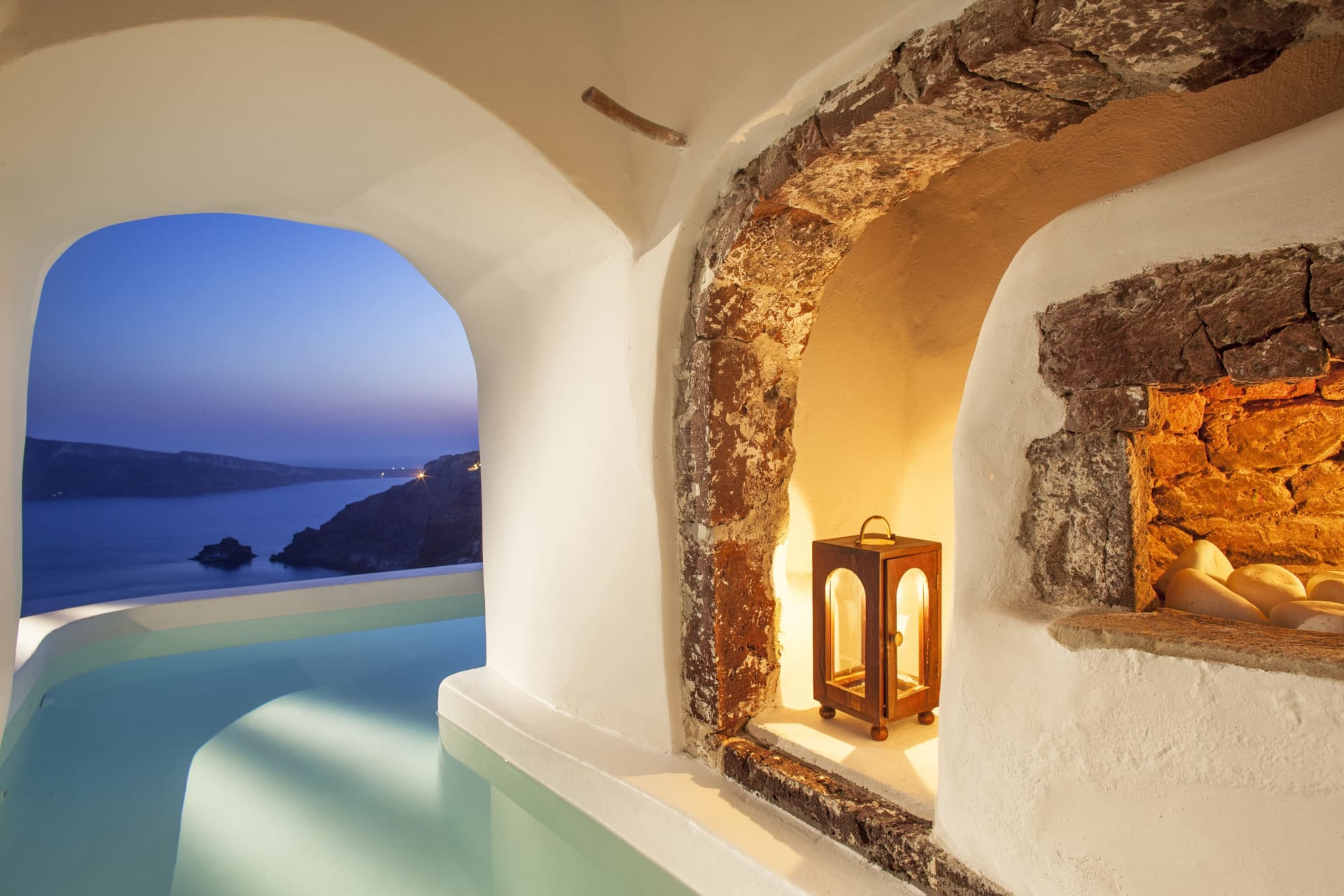 A stone fireplace lights a stone-walled room with a pool that extends outside and overlooks the Mediterranean