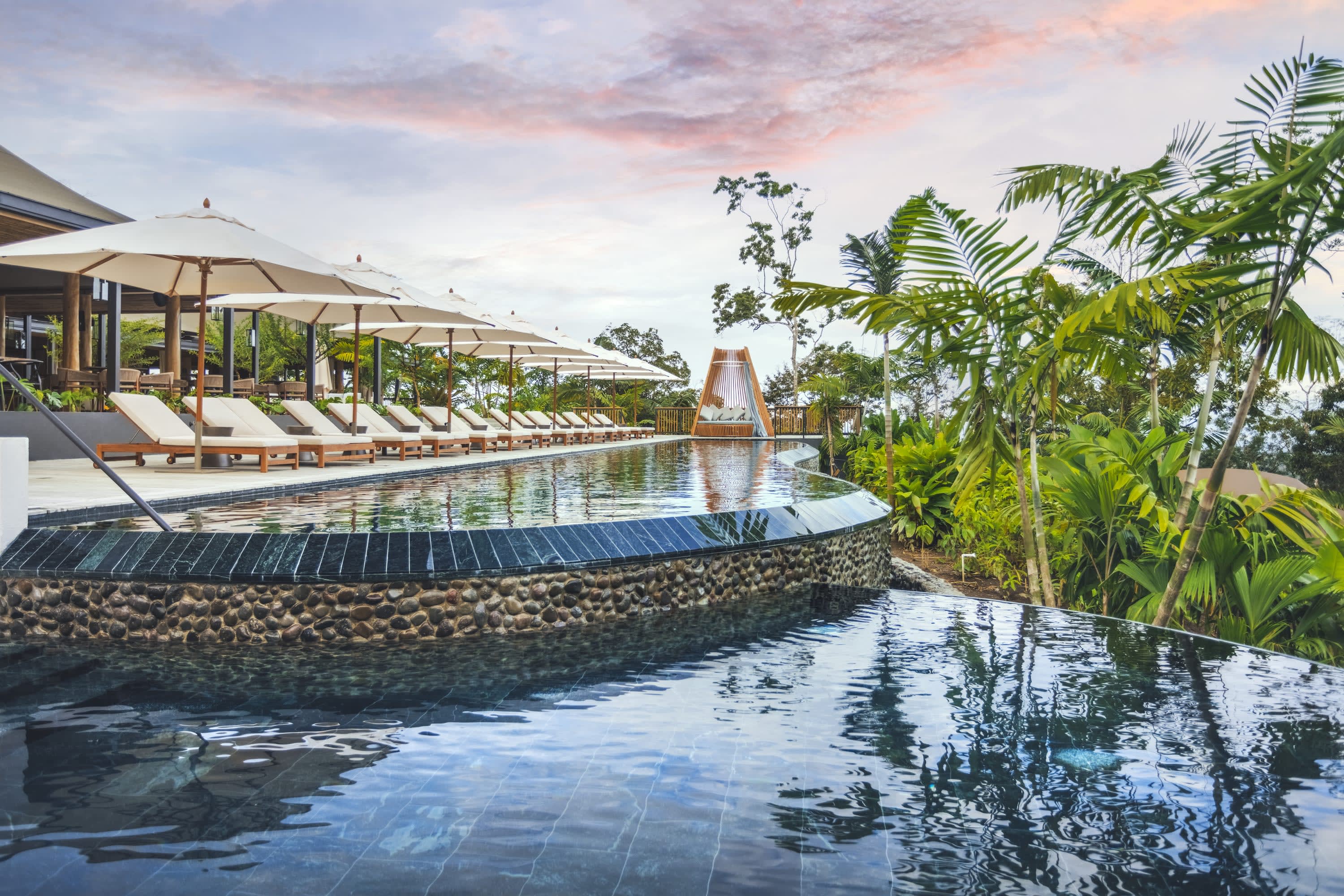 A chic pool area with luxe lounge chairs, jungle foliage and colorful clouds overhead
