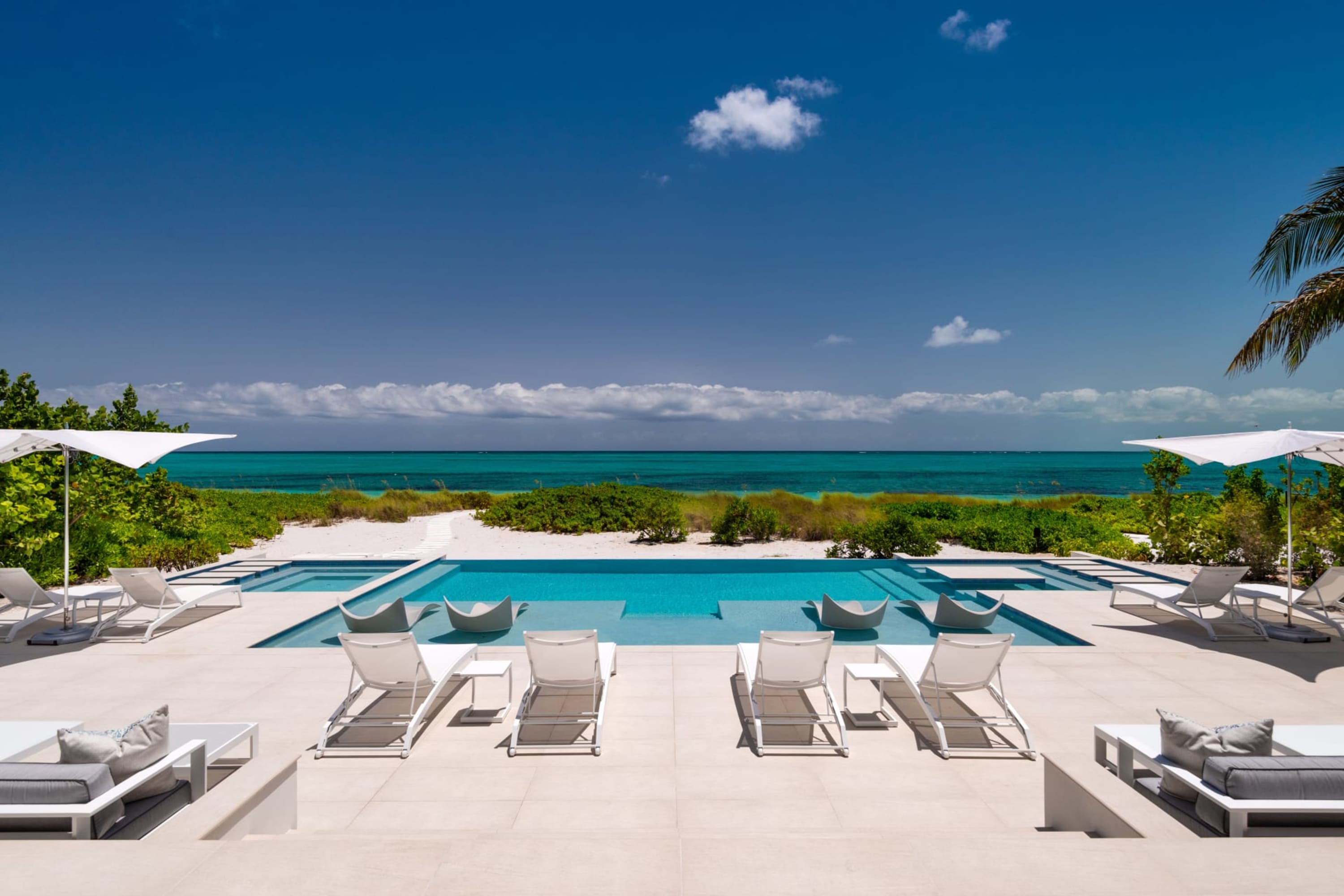 Chic lounge chairs sit before a turquoise pool, beach vegetation and the ocean