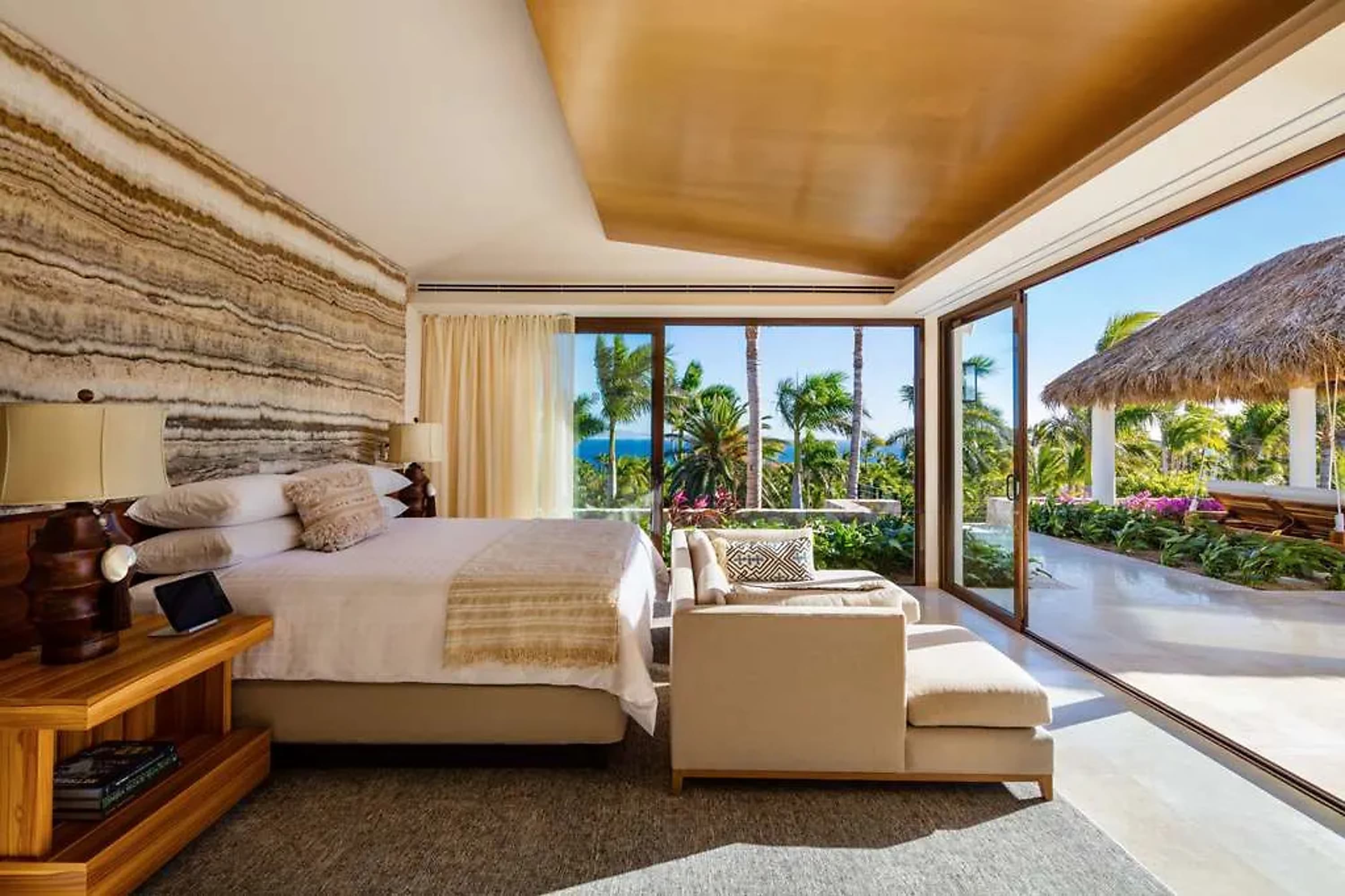 Spacious suite with plush bedding, tropical but chic decor and floor-to-ceiling windows and sliding doors revealing thatch-roofed hotel buildings