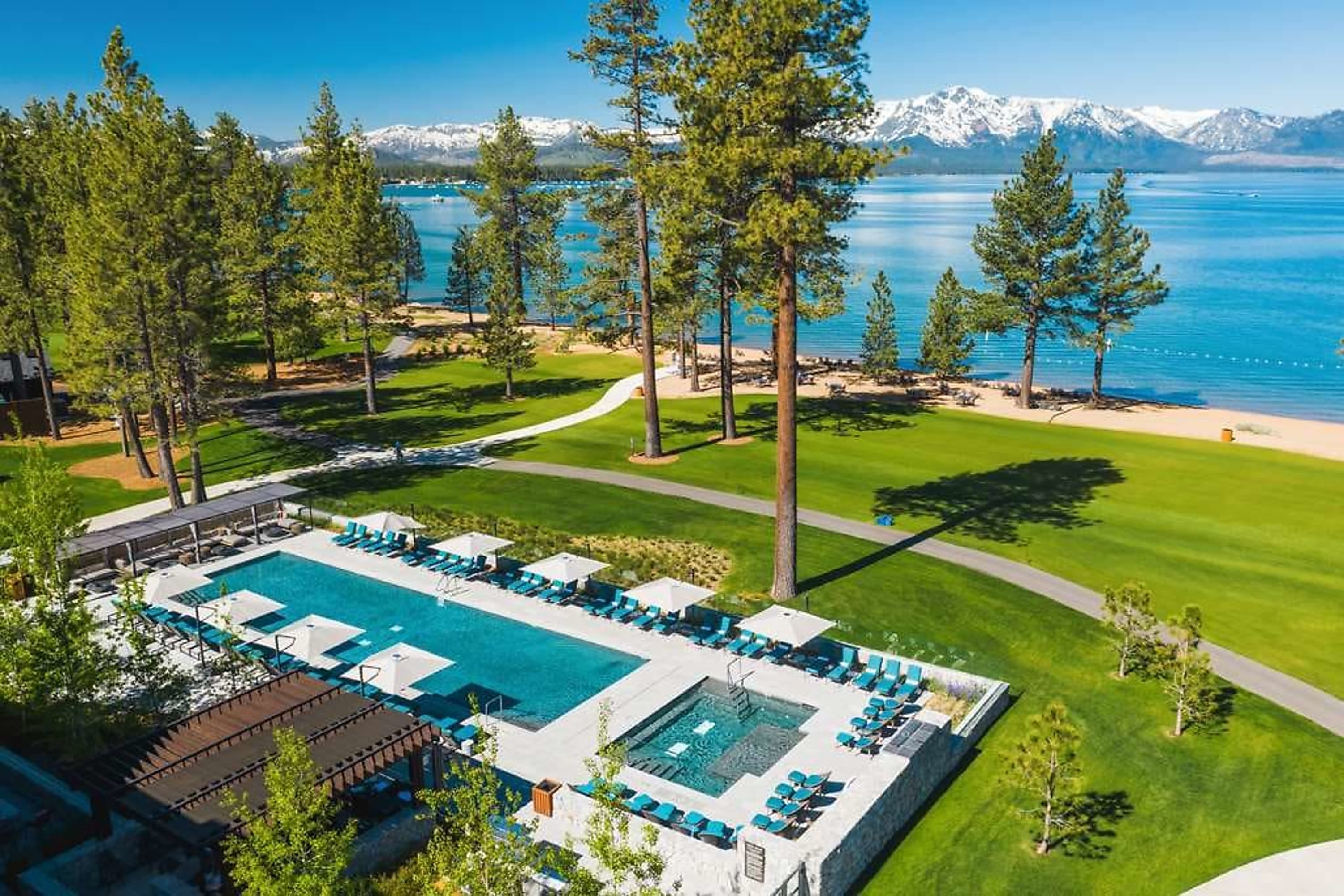 Chic pool area next to manicured resort grounds. In the distance a lakeside beach borders sapphire-blue water with snowcapped mountains in the far distance