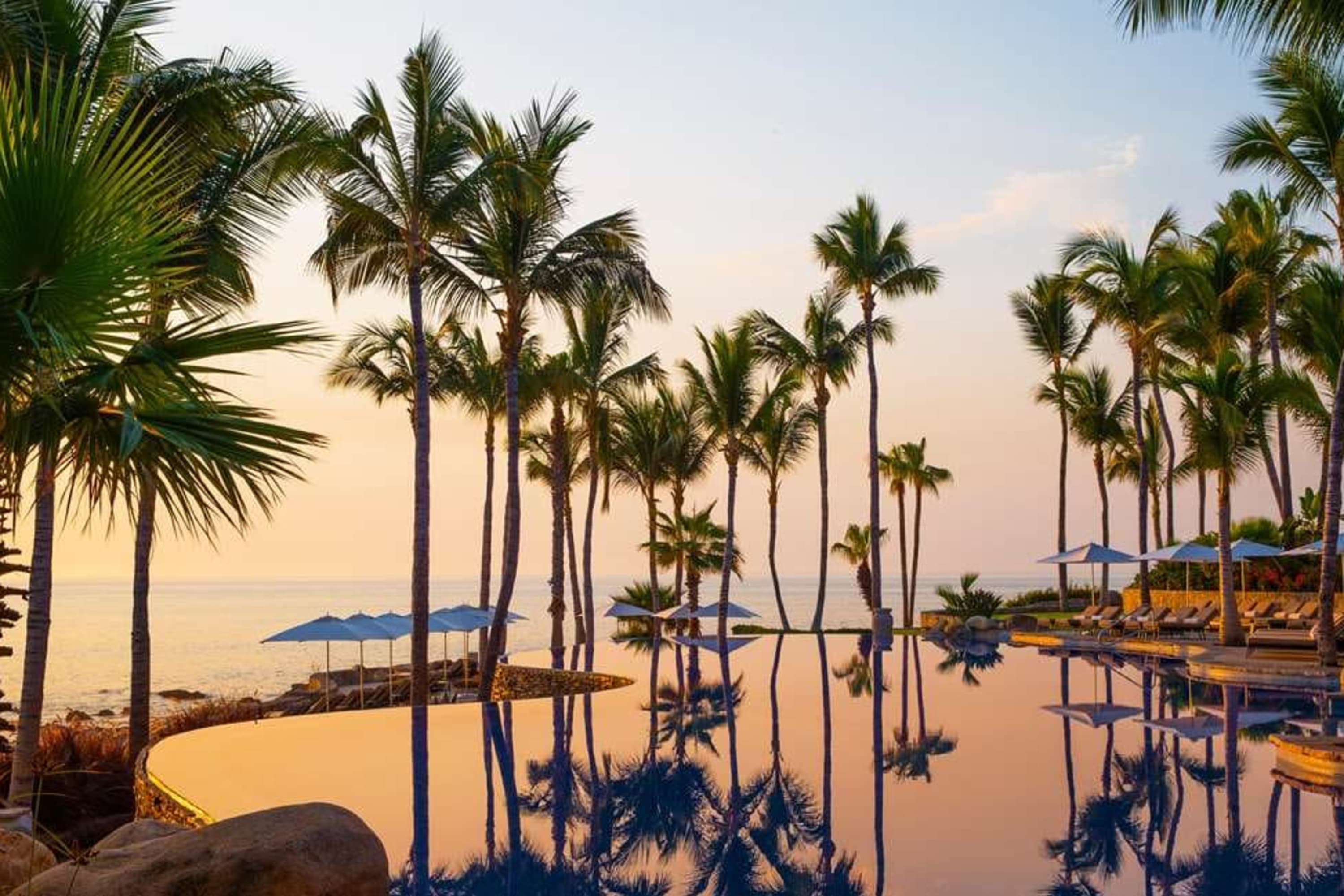 At sunset: palm trees reflect off a chic pool with beach loungers and umbrellas on the far side