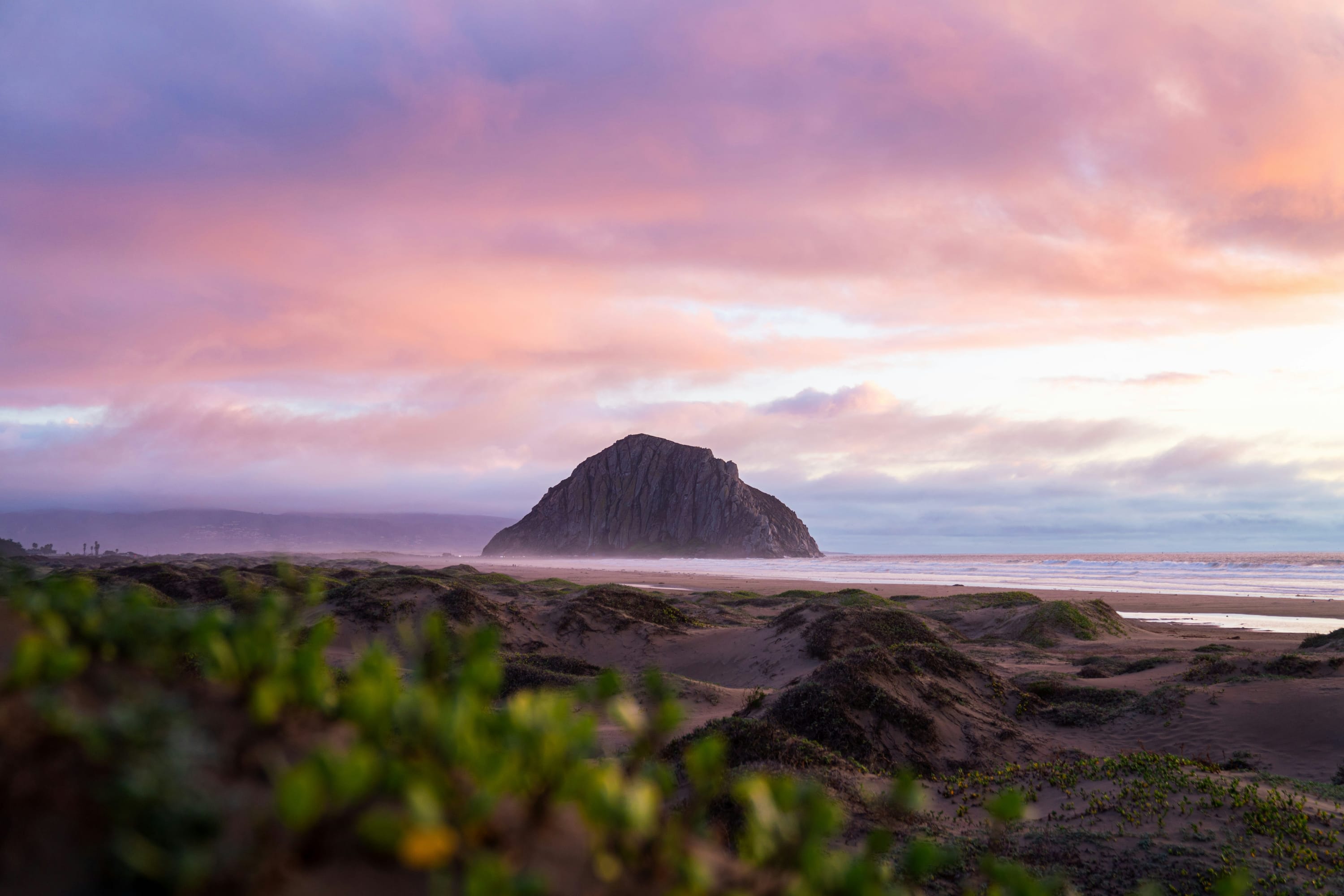 View from a quiet, empty beach: The iconic Morro Bay Rock stands in the distance under a pink sky