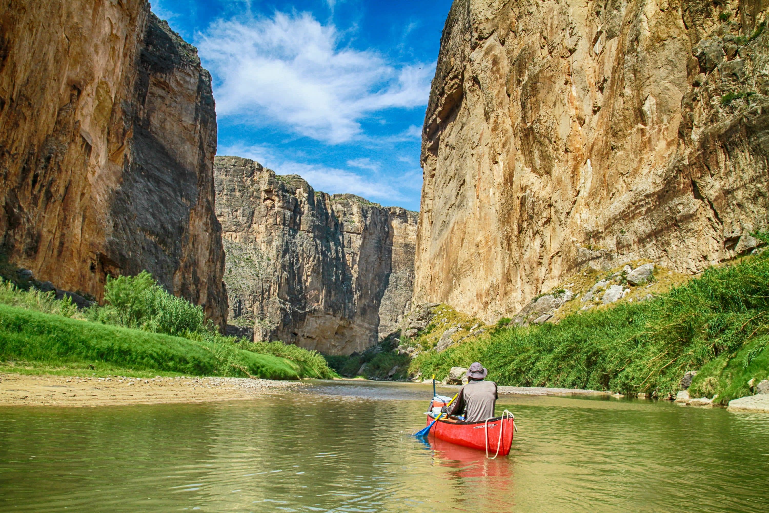 A guide in a red kayak paddles through a dramatic canyon, with lush vegetation on either side of the narrow river