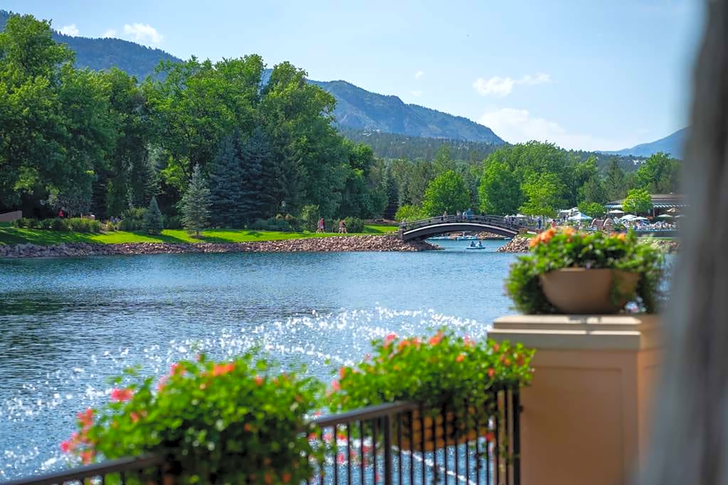 Out-of-focus flower baskets sit on a wrought iron railing before a calm lake surrounded by lush trees