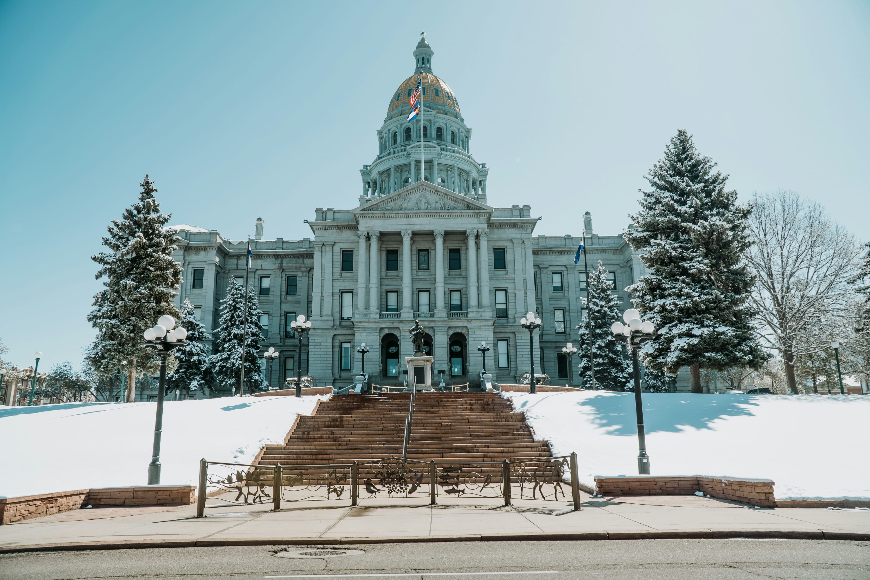 Clean street leads up to snow-covered hills and trees before the 19th-century architecture of the Denver Capitol Building on a sunny day