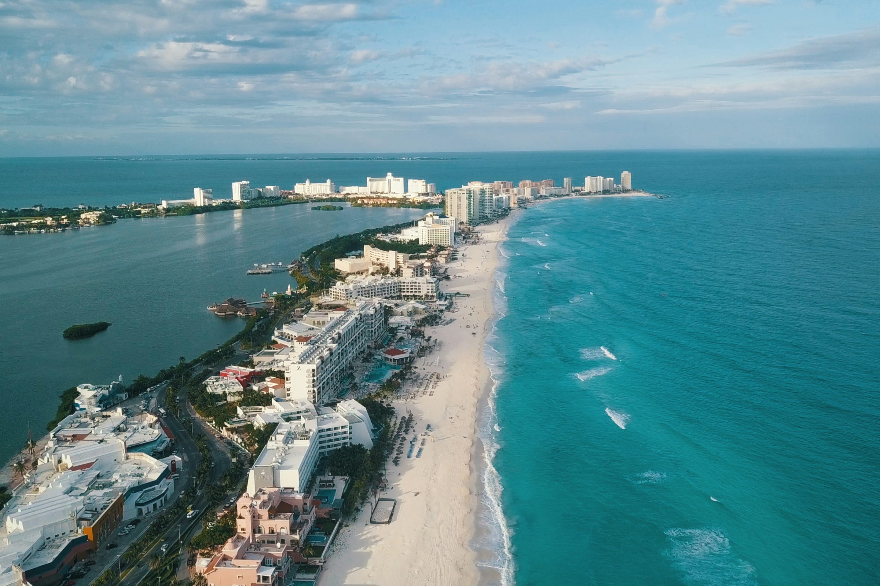 Aerial view of a long strip of land surrounded on either side by sapphire-blue water and filled with luxury resorts. Some clouds provide a touch of shade