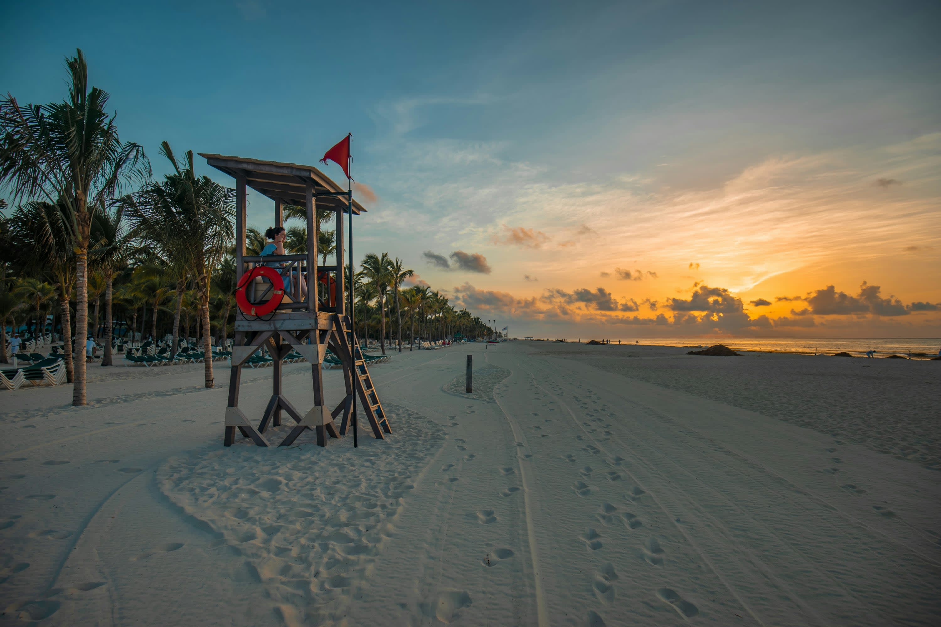 A lifeguard sits in her tower on a largely empty beach watching the sunrise over the ocean horizon