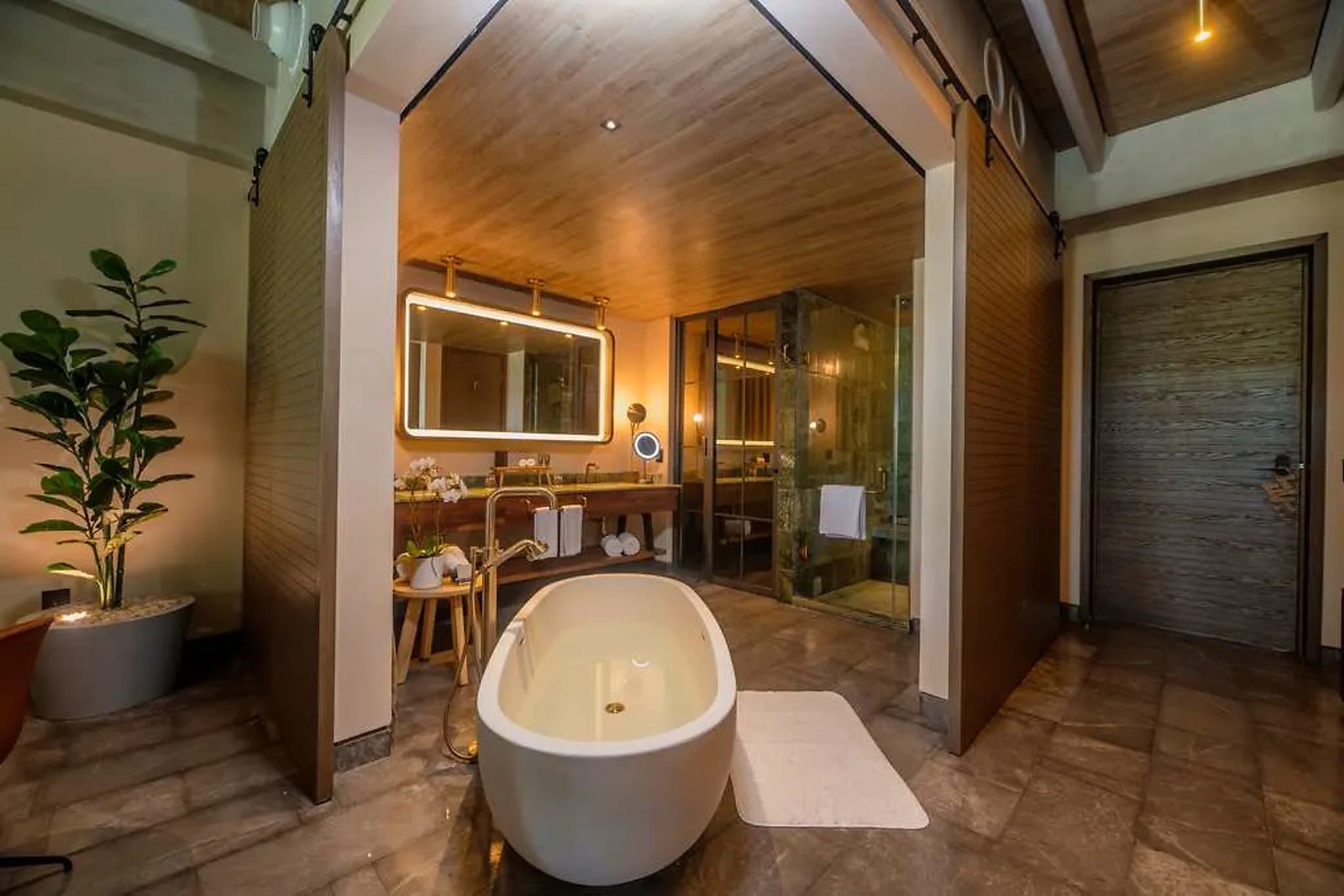 An expansive shower and deep-soaking tub area with hardwood floors, potted plants, gold metal accents and rollaway doors for privacy