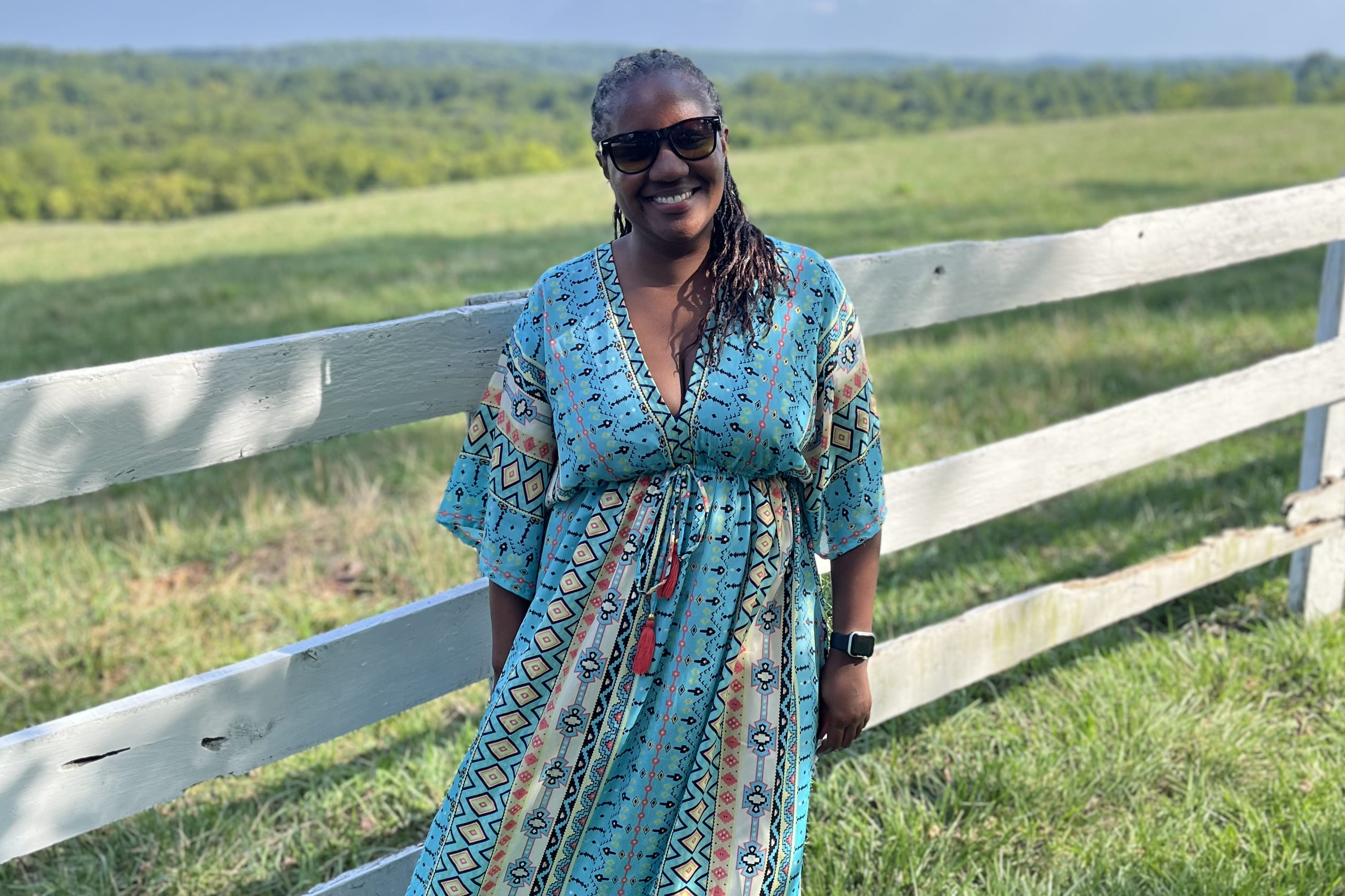 woman standing in front a white fence in a meadow with blue skies behind