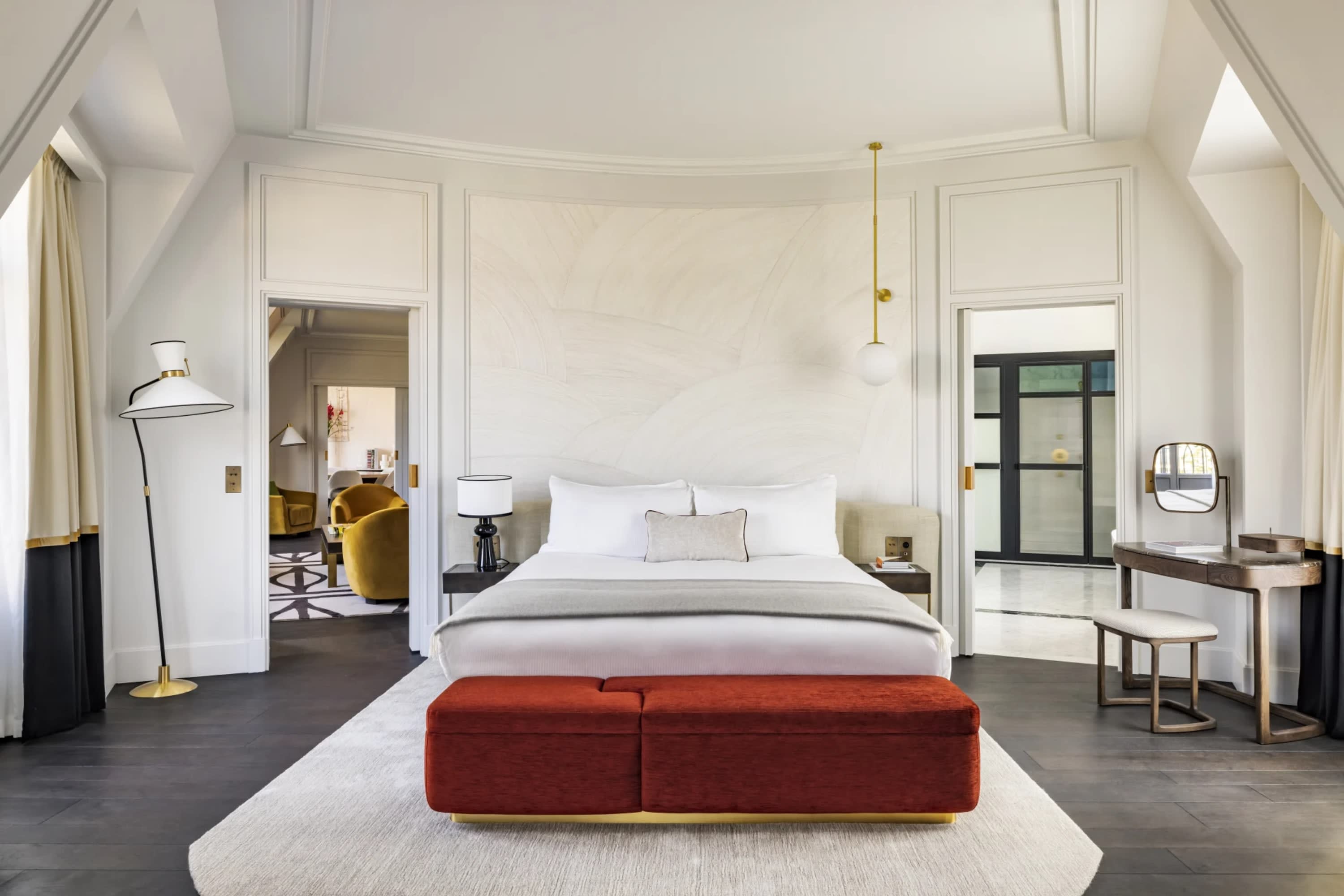 Whitewashed furnishings contrast with dark wood floors and a bold red ottoman in a spacious hotel room
