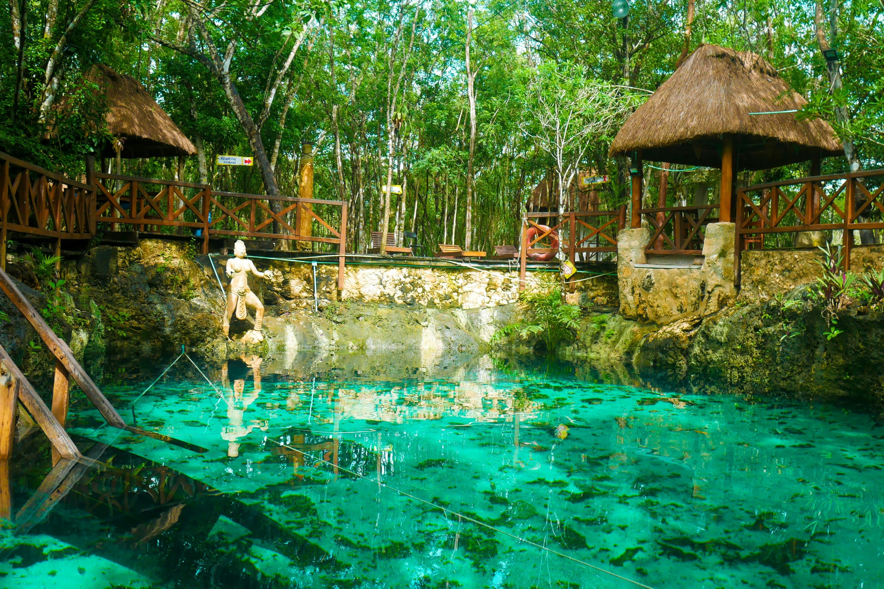 Turquoise water with a ladder descending into it and small rock border surrounding the body. A small Mayan statue stands at the opposite end before wooden railings and tropical jungle