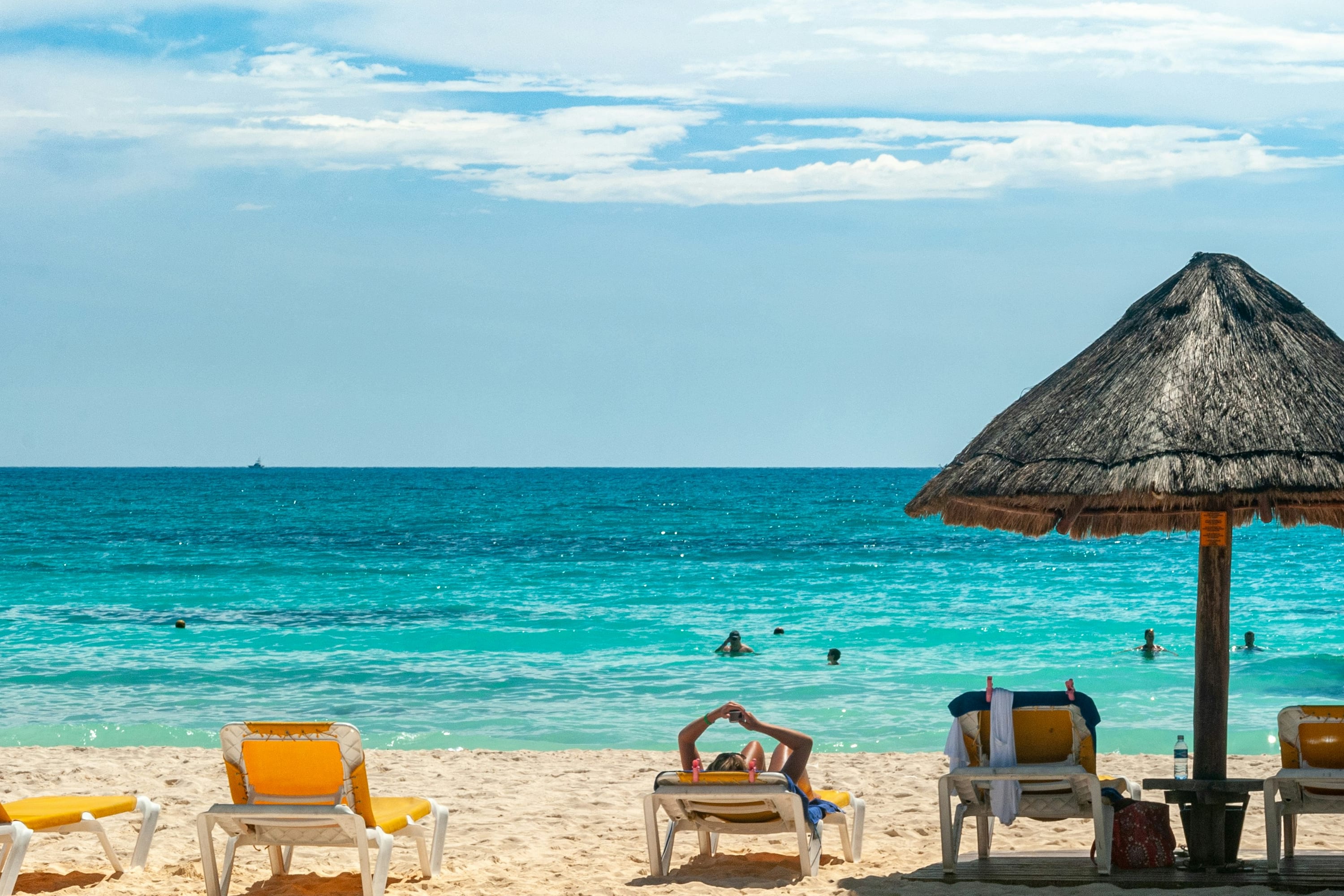 A sunny beach with a handful of people in the water or on loungers under a thatch umbrella