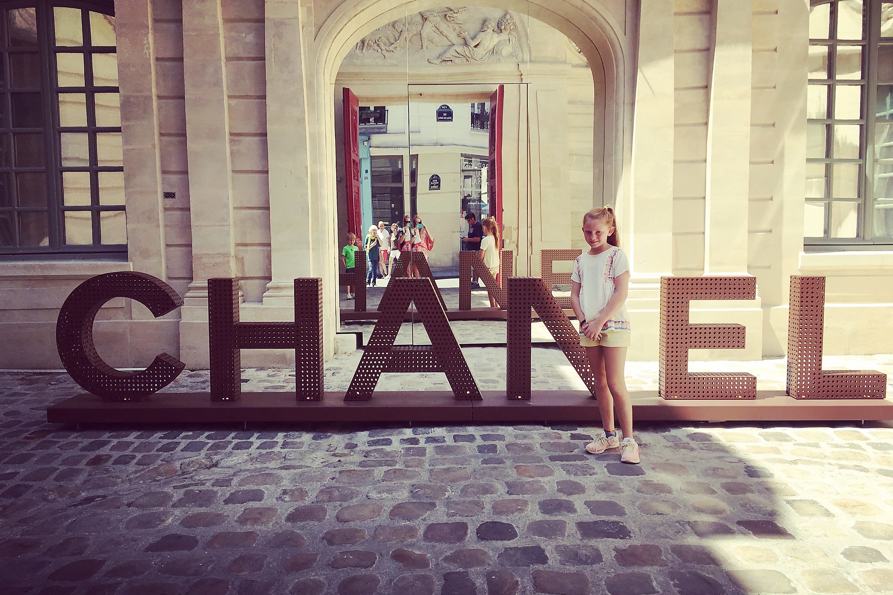 A young girl stands next to a 3D sign reading "Chanel" in a historic part of Paris with cobblestone streets