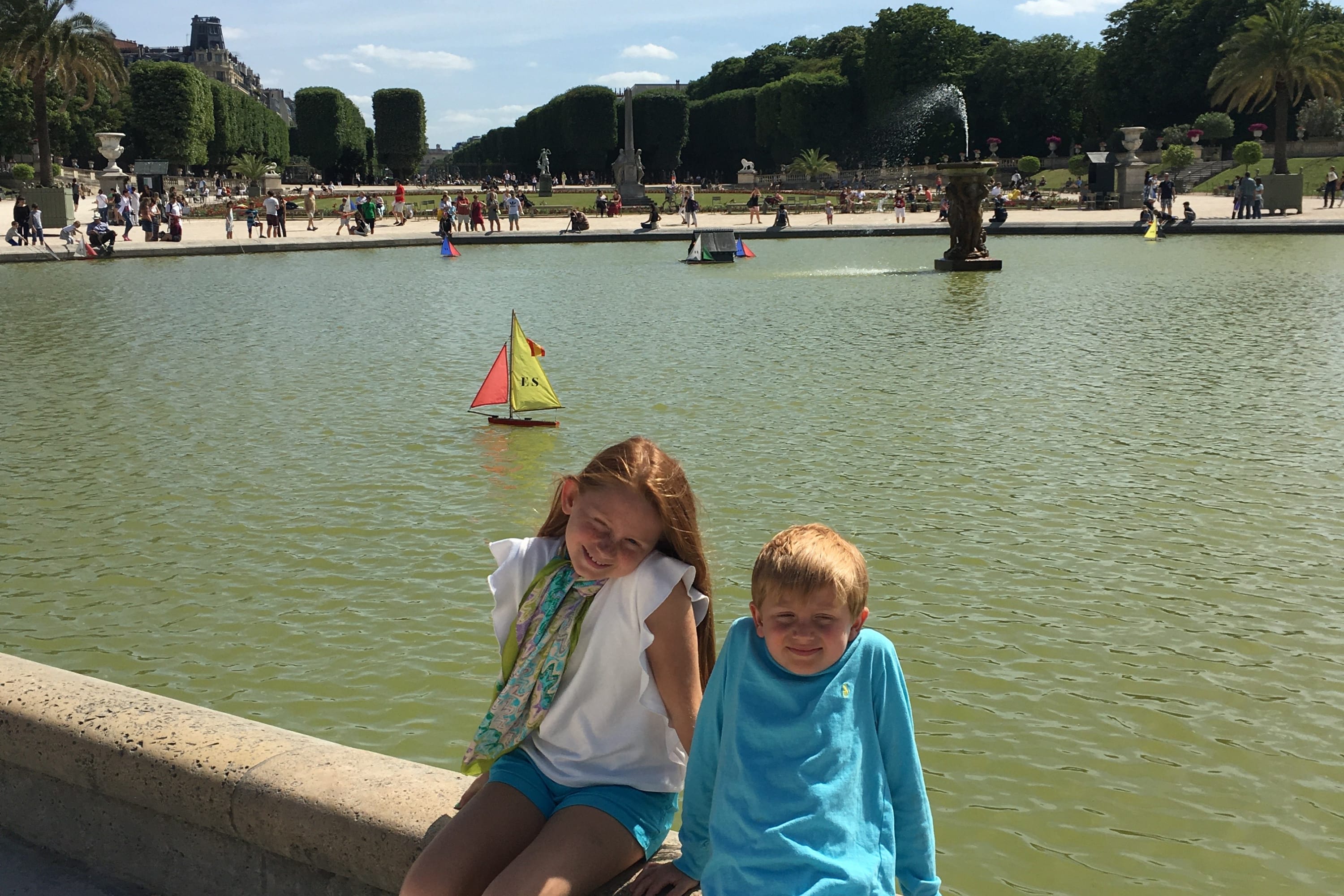 Two young children sit on a ledge in front of a large pond in a Parisian park