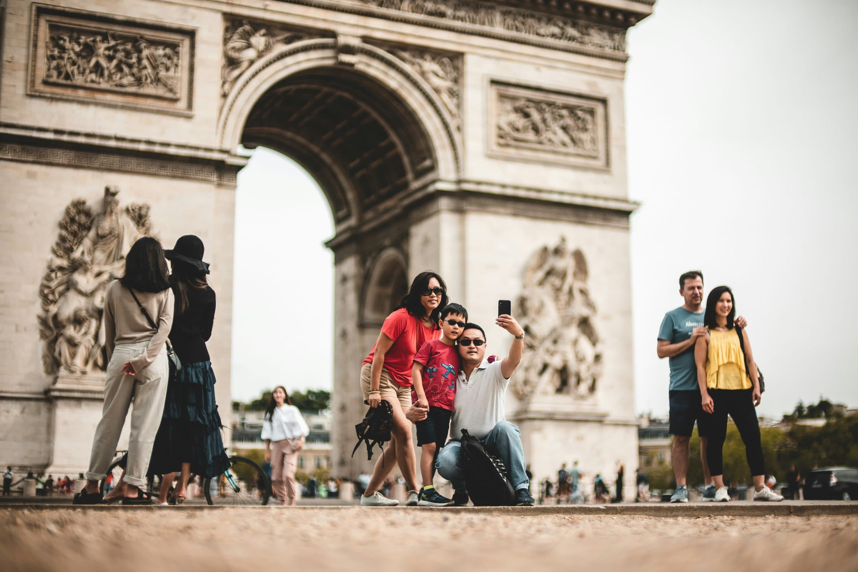 A mom and a dad pose with their child for a selfie in front of Paris' Arc de Triomphe