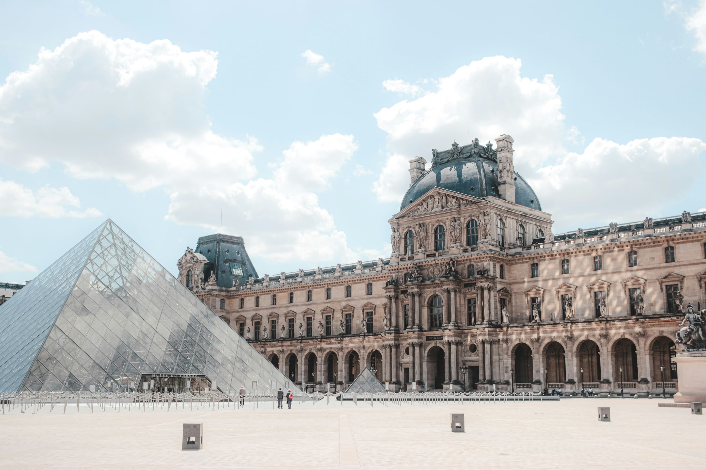 A diagonal view of the Louvre, with the ornate historic structure in the background and the glass pyramid out in front, partially out of view