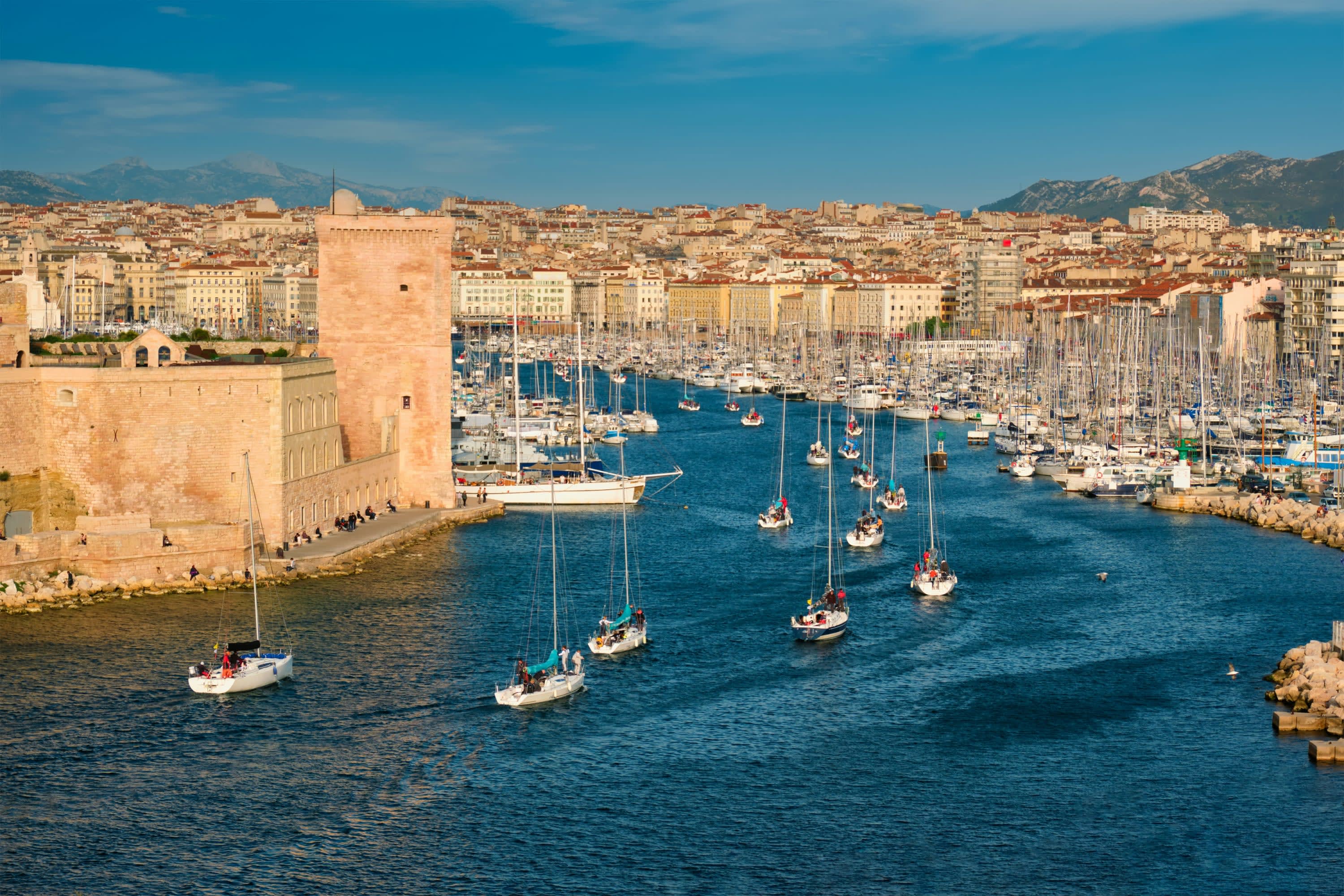 A stream of sailboats enter Marseille harbor, passing historic forts and other buildings