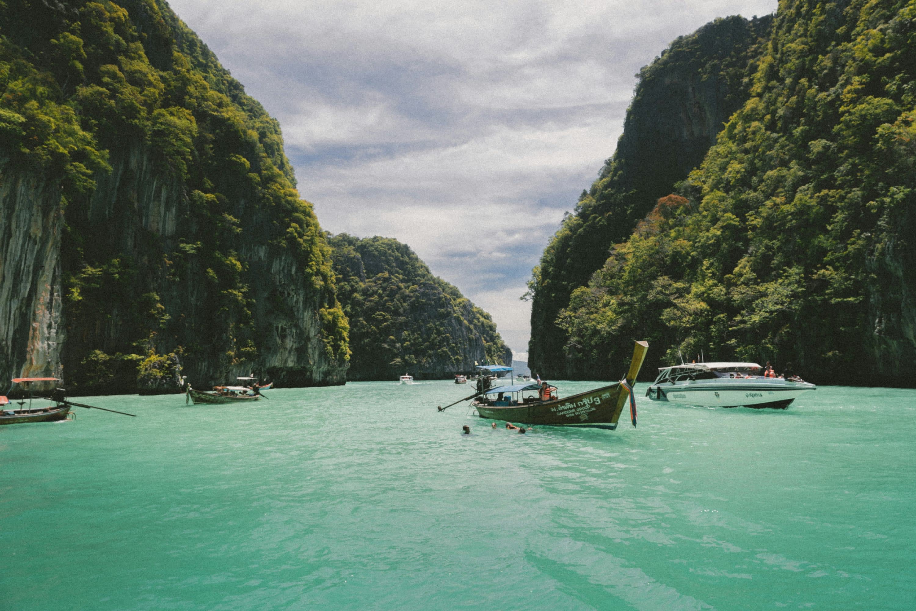 A mix of modern and traditional boats sail through a tropical river valley while a few people swim in the turquoise water