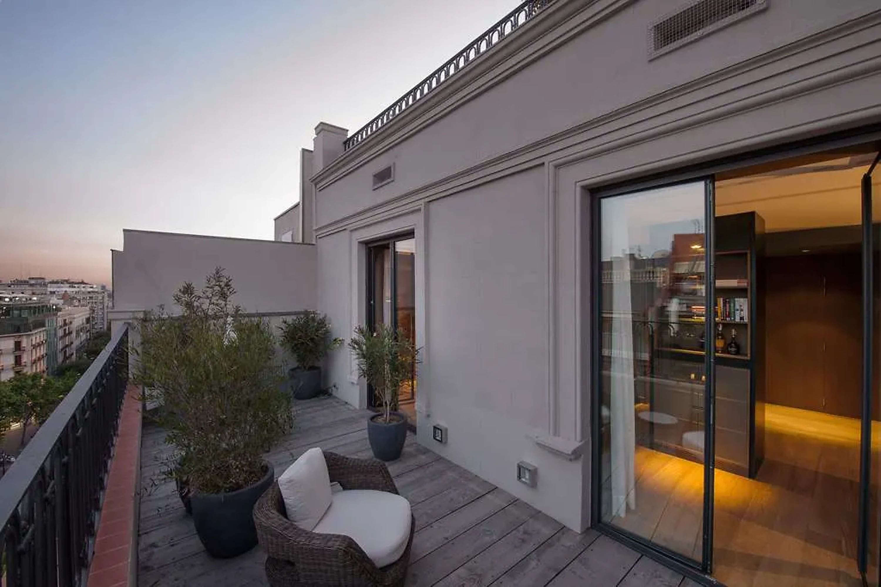 Chic balcony with potted plants and a plush outdoor chair. A moody room with hardwood floors is visible through glass doors