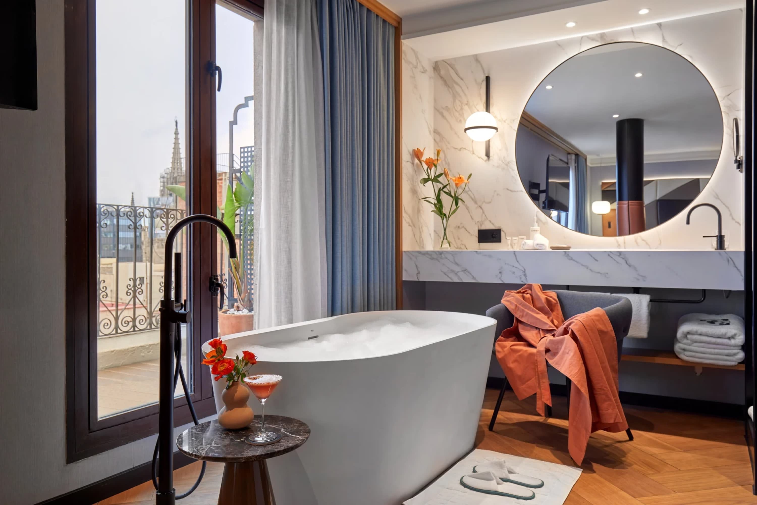 A deep soaking tub filled with bubbles in a massive bathroom stands next to floor-to-ceiling window revealing Barcelona's historic skyline