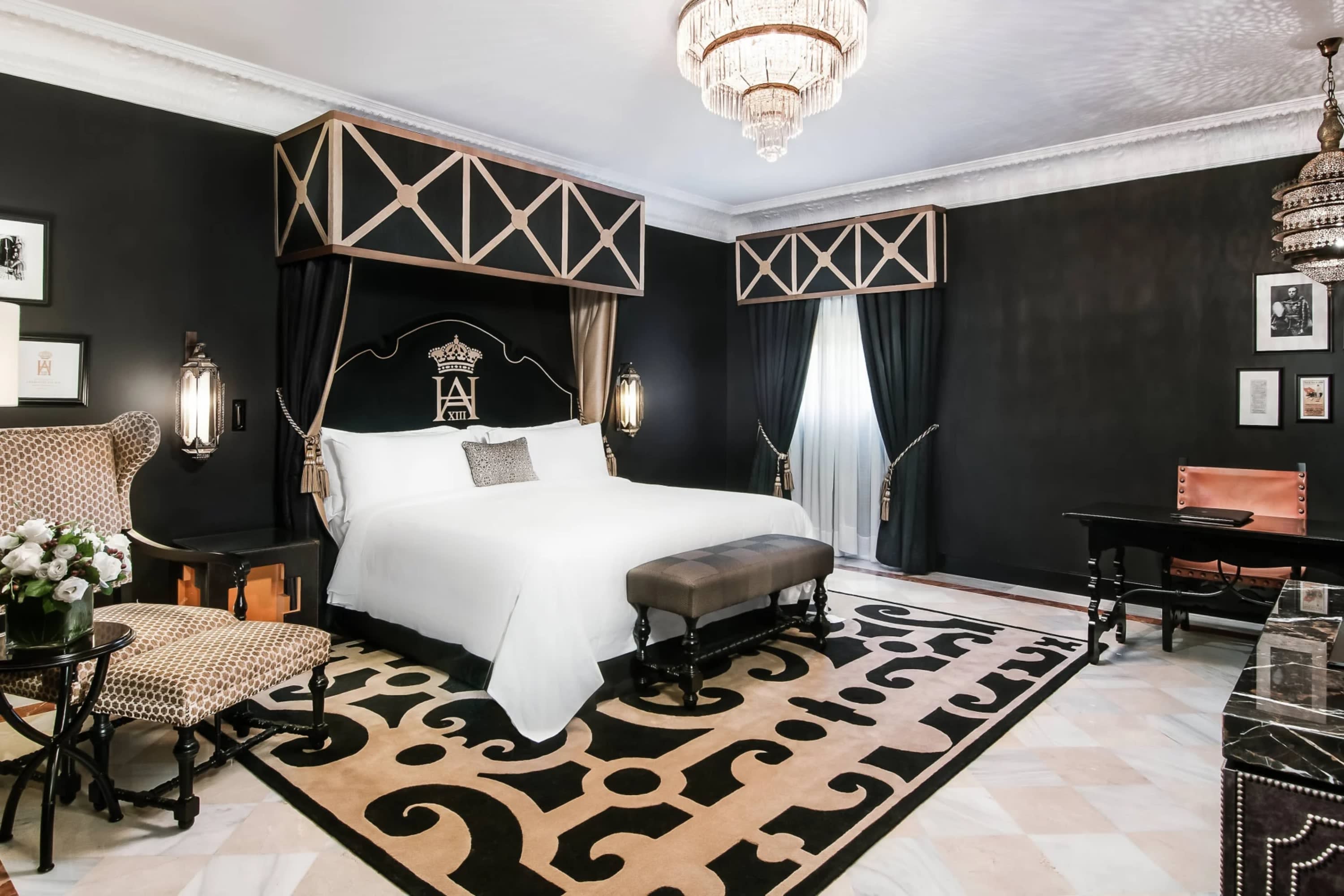 Exquisite hotel room with classical aesthetic: thick black drapes on all walls, a massive bed, ornate rug, glass chandelier and more.