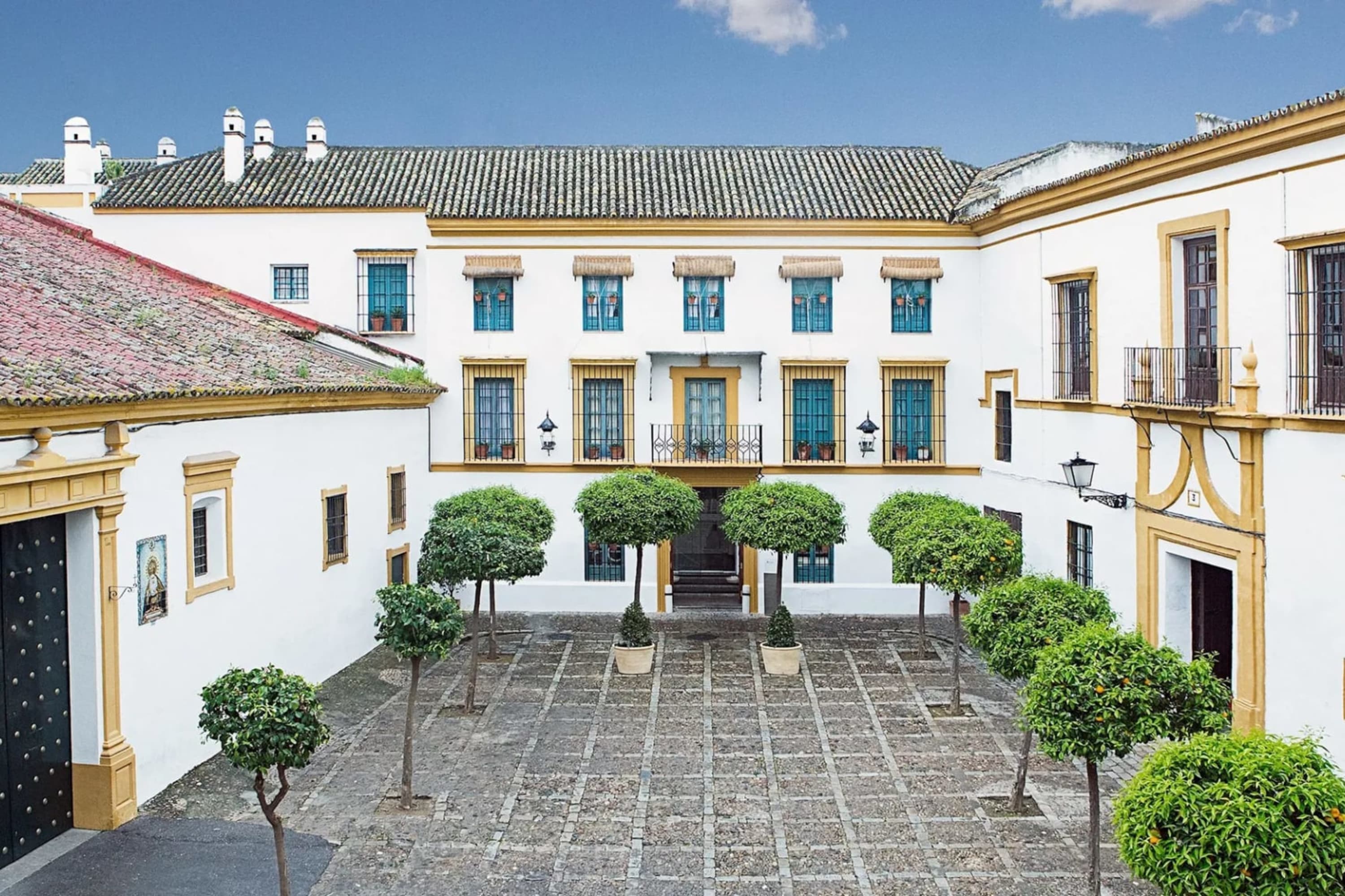 Historic, tree-line courtyard against whitewashed walls with wrought iron details, blue shutters and Spanish roof tiles