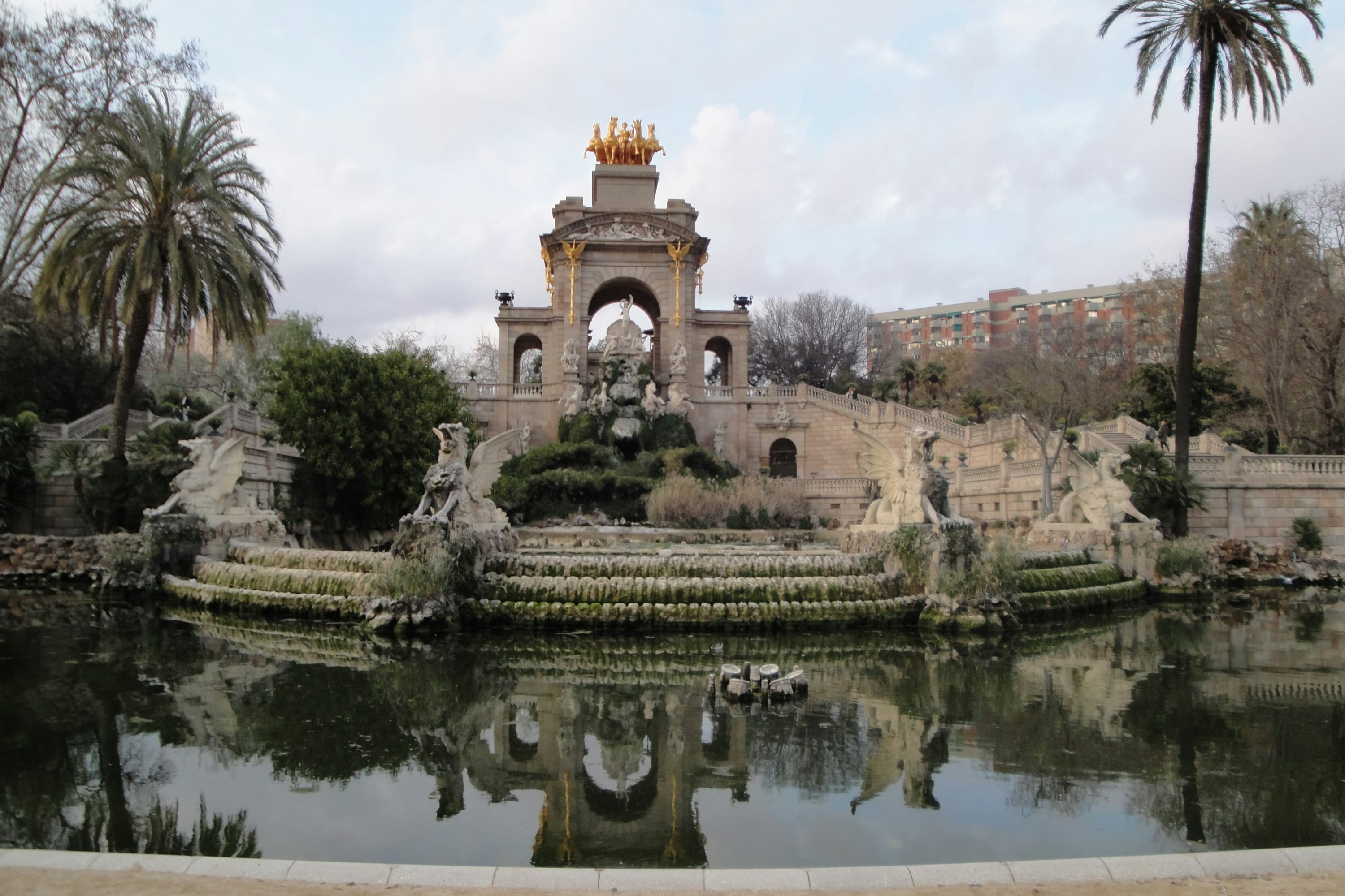 Neoclassical fountain surrounded by a small pond and palm trees on a cloudy day