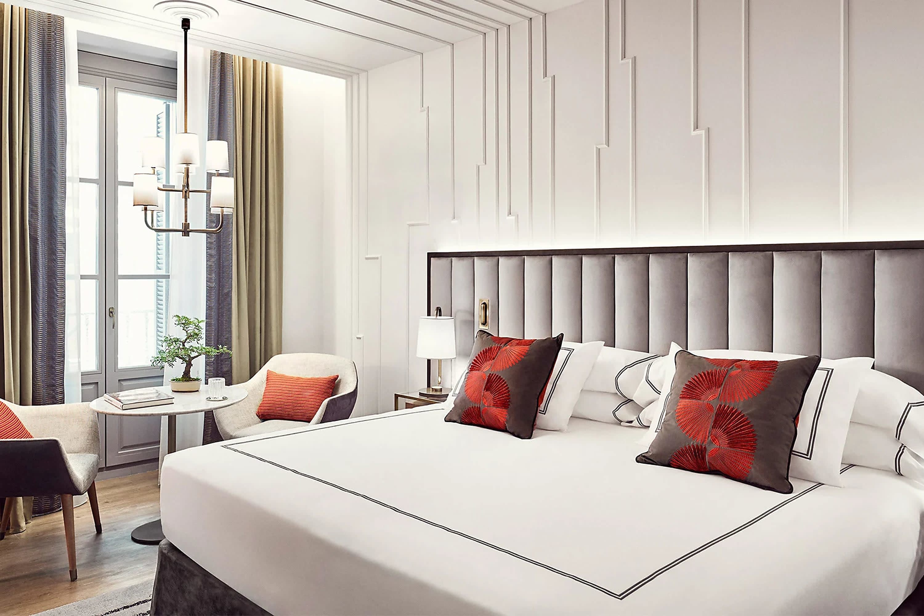 Chic hotel room with intricate wall details, an elegant light fixture over a small table and chairs and bold red pillows on a white-and-black bedspread