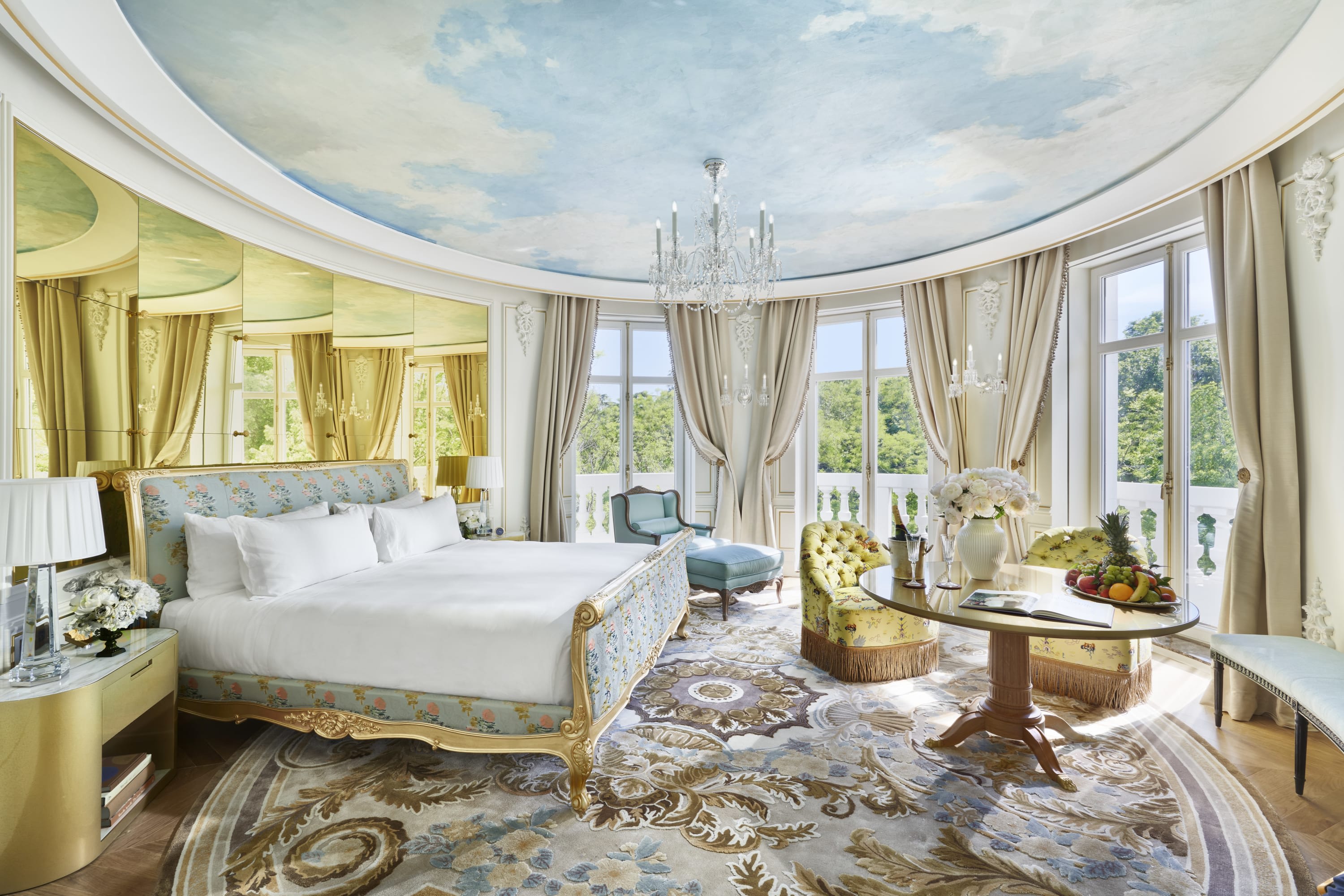 Elite hotel room with a circular layout, gilded curtains, an opulent bedframe and plush bedding, an intricate high-end rug, gilded mirrors and a sky mural on the ceiling