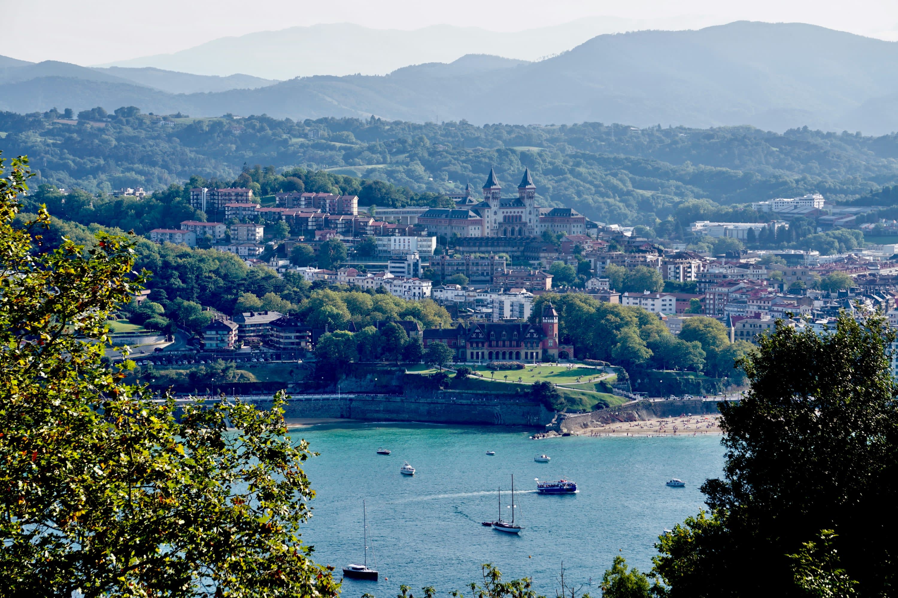 Framed by trees: a view across a body of water filled with sailboats of historic architecture built into a hillside with misty mountains in the distance