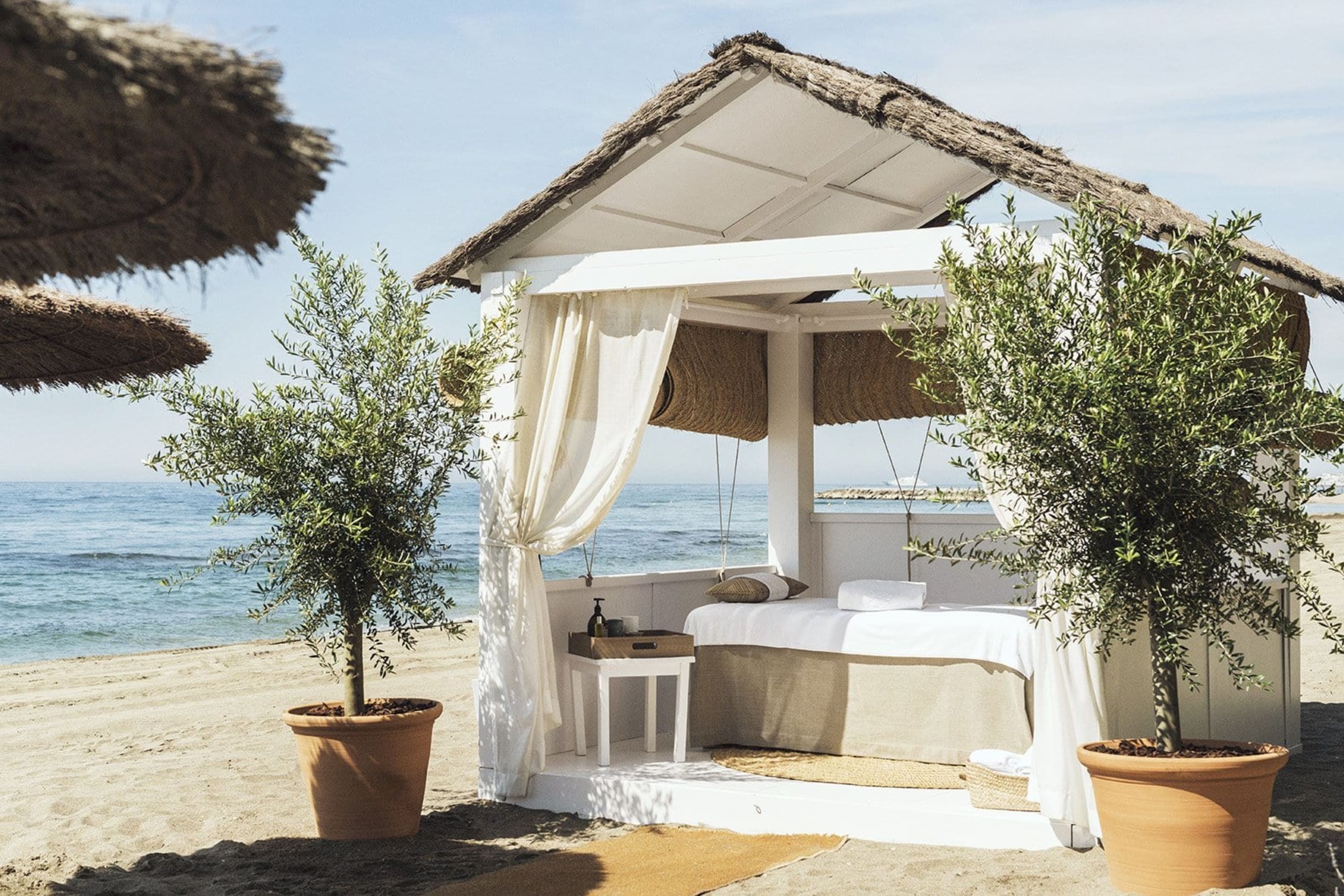 A dreamy beach canopy with potted plants, white curtains, a shaded day bed and the Mediterranean Sea in the background