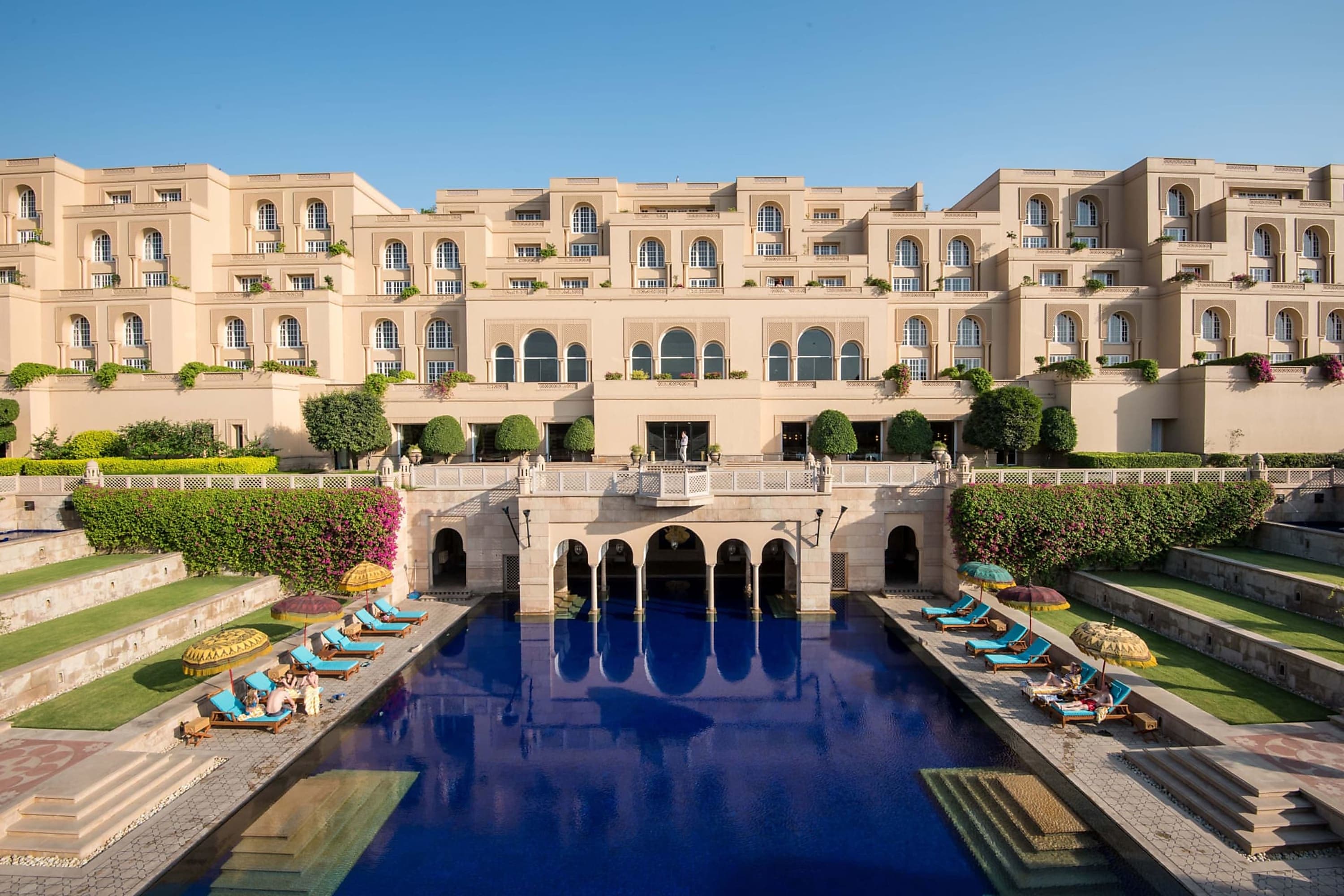 Long courtyard pool leads to a palace-like hotel. On either side of the pool, stone stairs with lawns rise